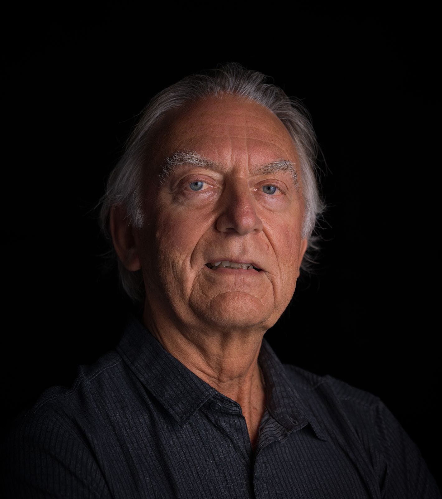 An older man with gray hair and blue eyes wearing a dark shirt, against a black background.