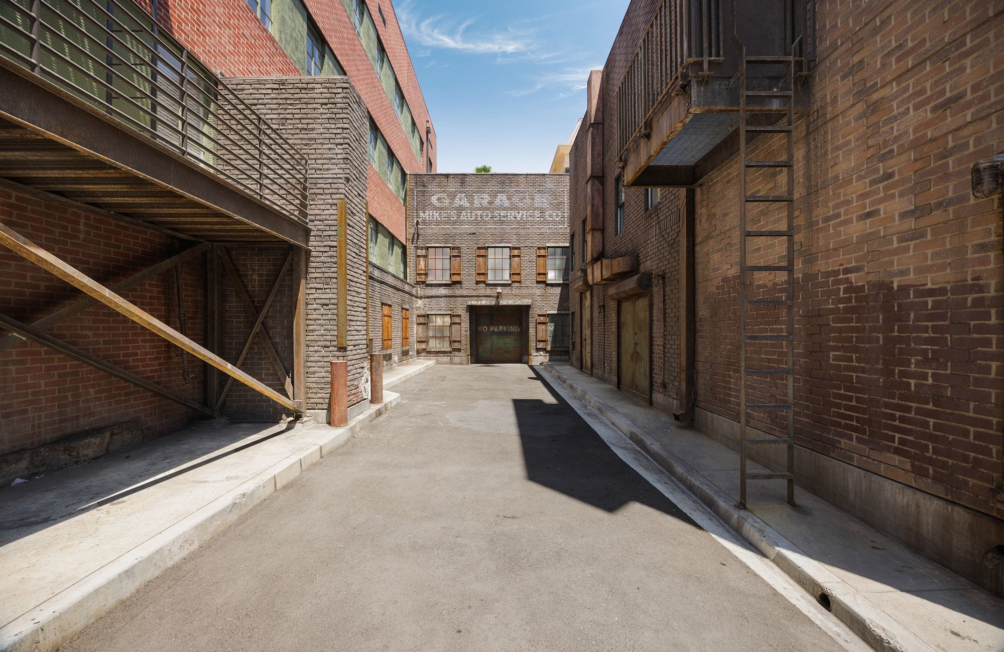 An urban alleyway between brick buildings with an elevated walkway and a fire escape ladder, leading to a garage sign.