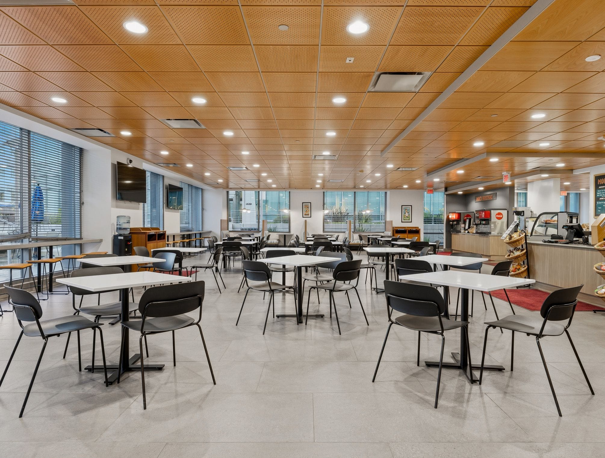 An empty, modern cafeteria with white tables, black chairs, a wood ceiling, and a food service counter.