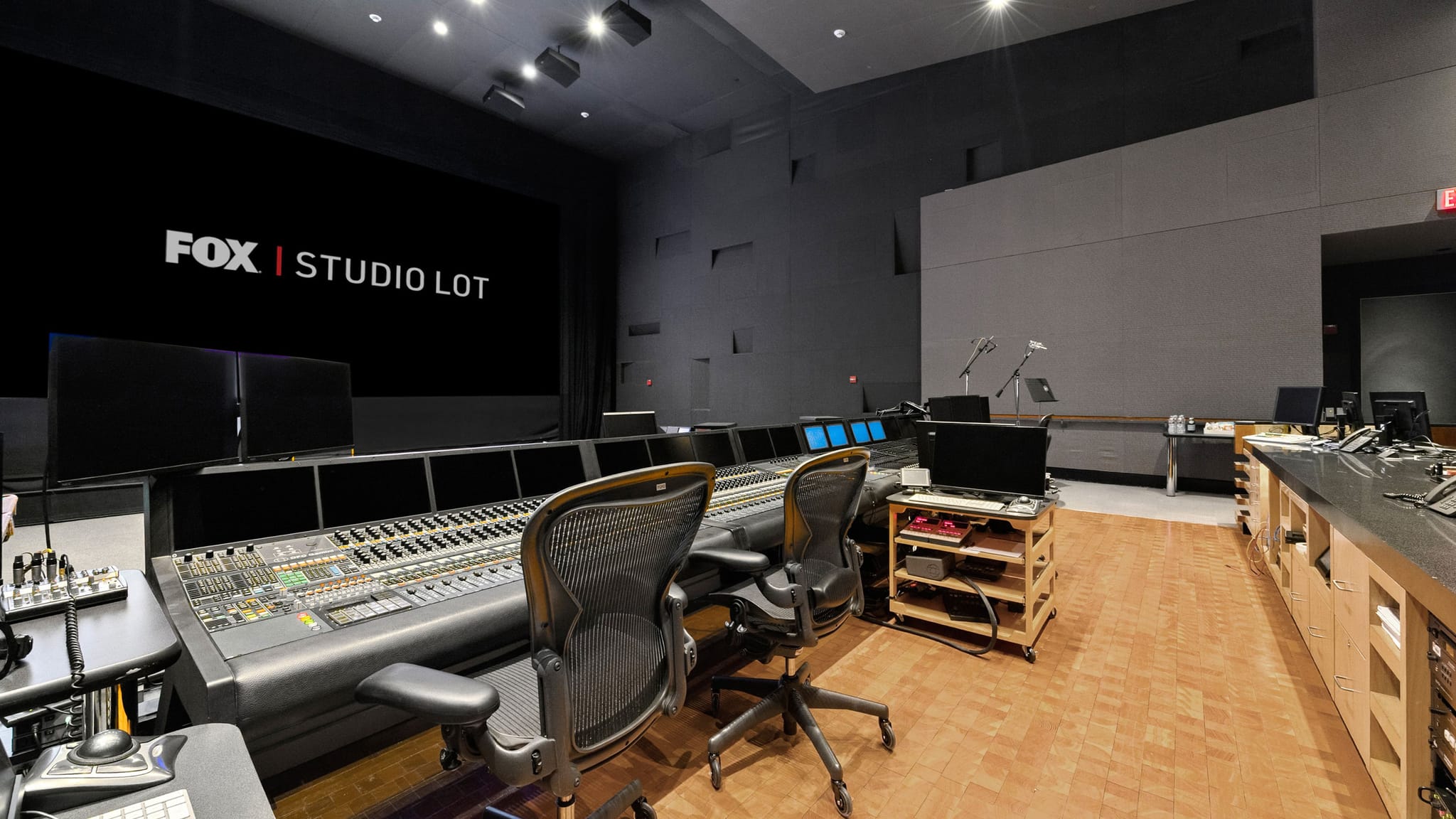 A modern studio control room with a large mixing console in the foreground and a screen displaying "FOX | STUDIO LOT" in the background.
