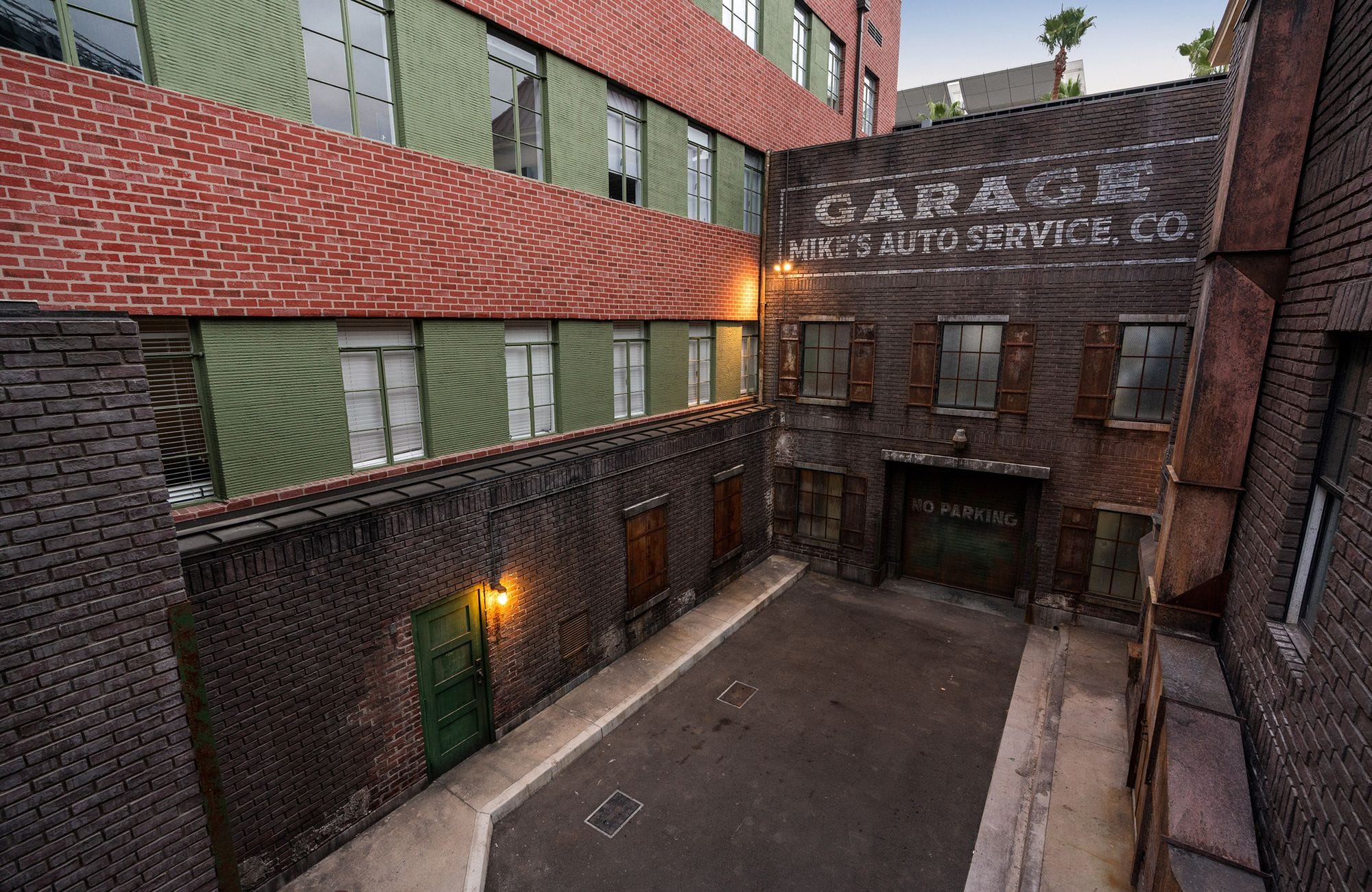 A brick alleyway with a vintage-style garage building featuring a faded "Mike's Auto Service" sign, flanked by buildings with green walls and red brick.