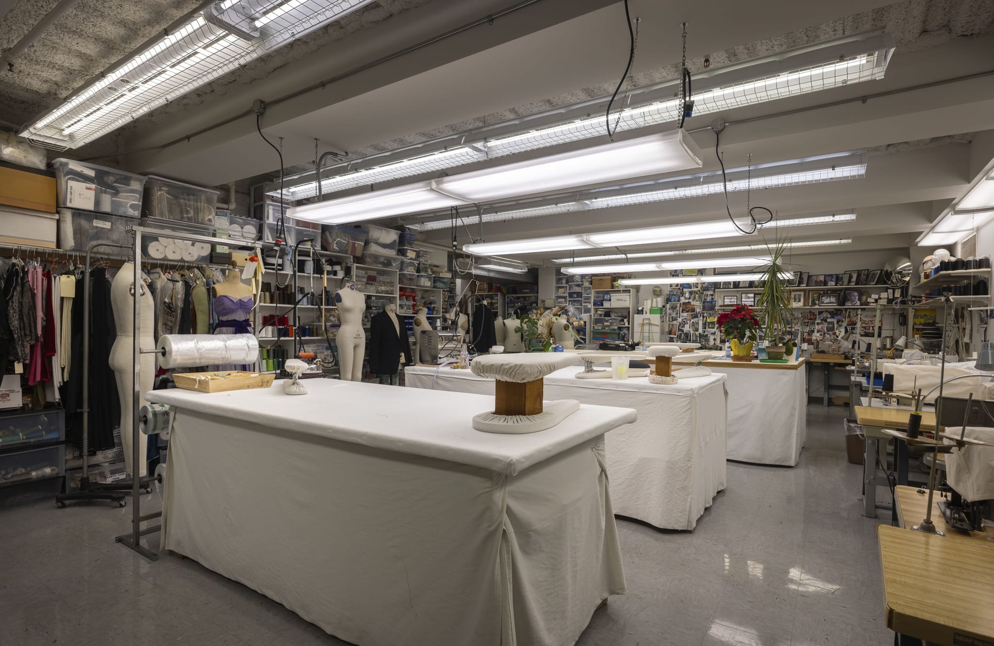 A brightly lit tailoring workshop with large white work tables, mannequins, clothing racks, and sewing machines.