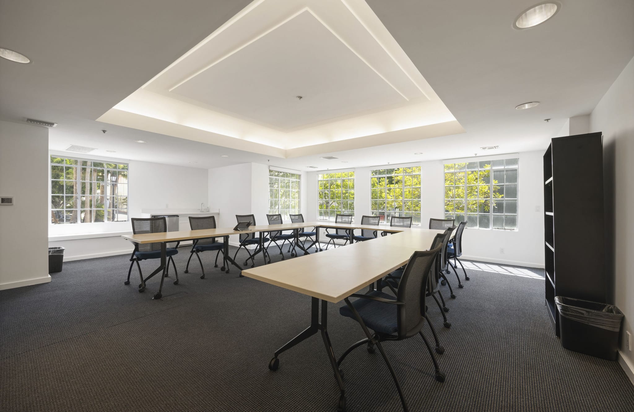 A bright meeting room with a U-shaped table, chairs, large windows, and a dark bookshelf.