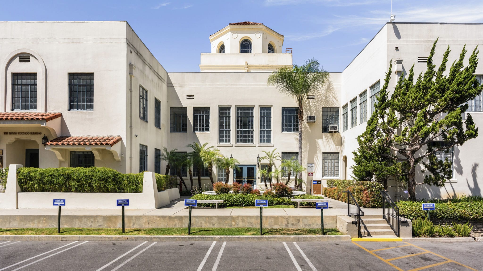 A light-colored, U-shaped building with a central courtyard and dome, seen from a parking lot on a sunny day.