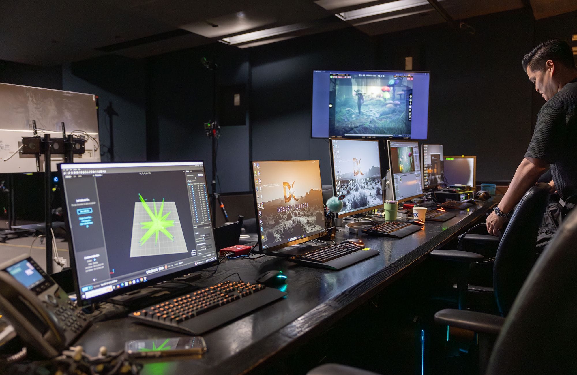 A person stands at a long desk with multiple computer monitors displaying game development software and video game footage.
