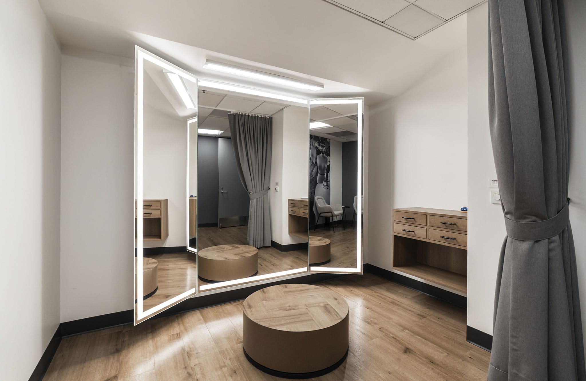 A modern fitting room with an illuminated three-panel mirror, two round ottomans, wooden console tables, and a grey curtain on a wooden floor.