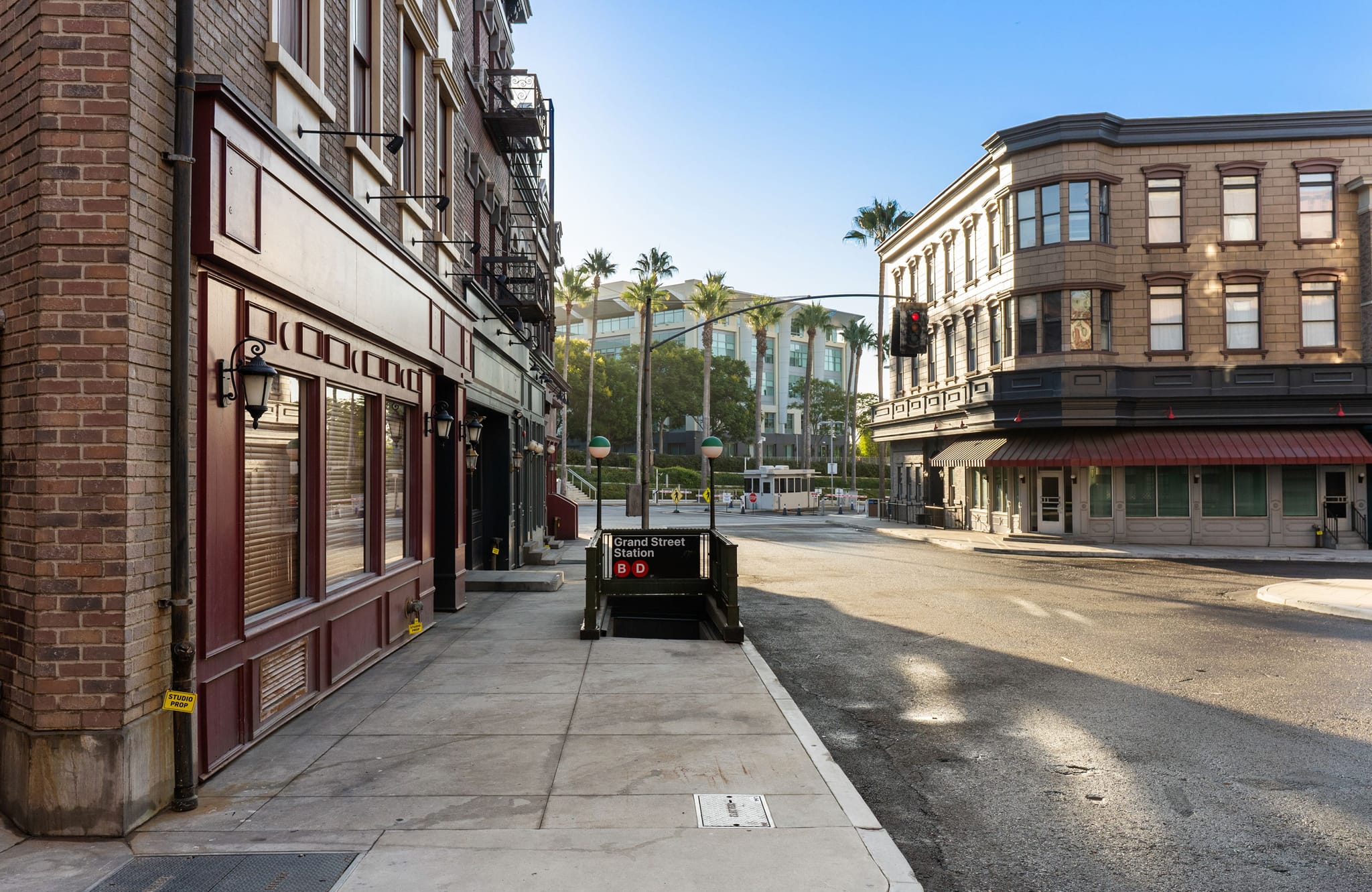 An empty street scene featuring brick buildings, a "Grand Street Station" subway entrance, and palm trees in the background.
