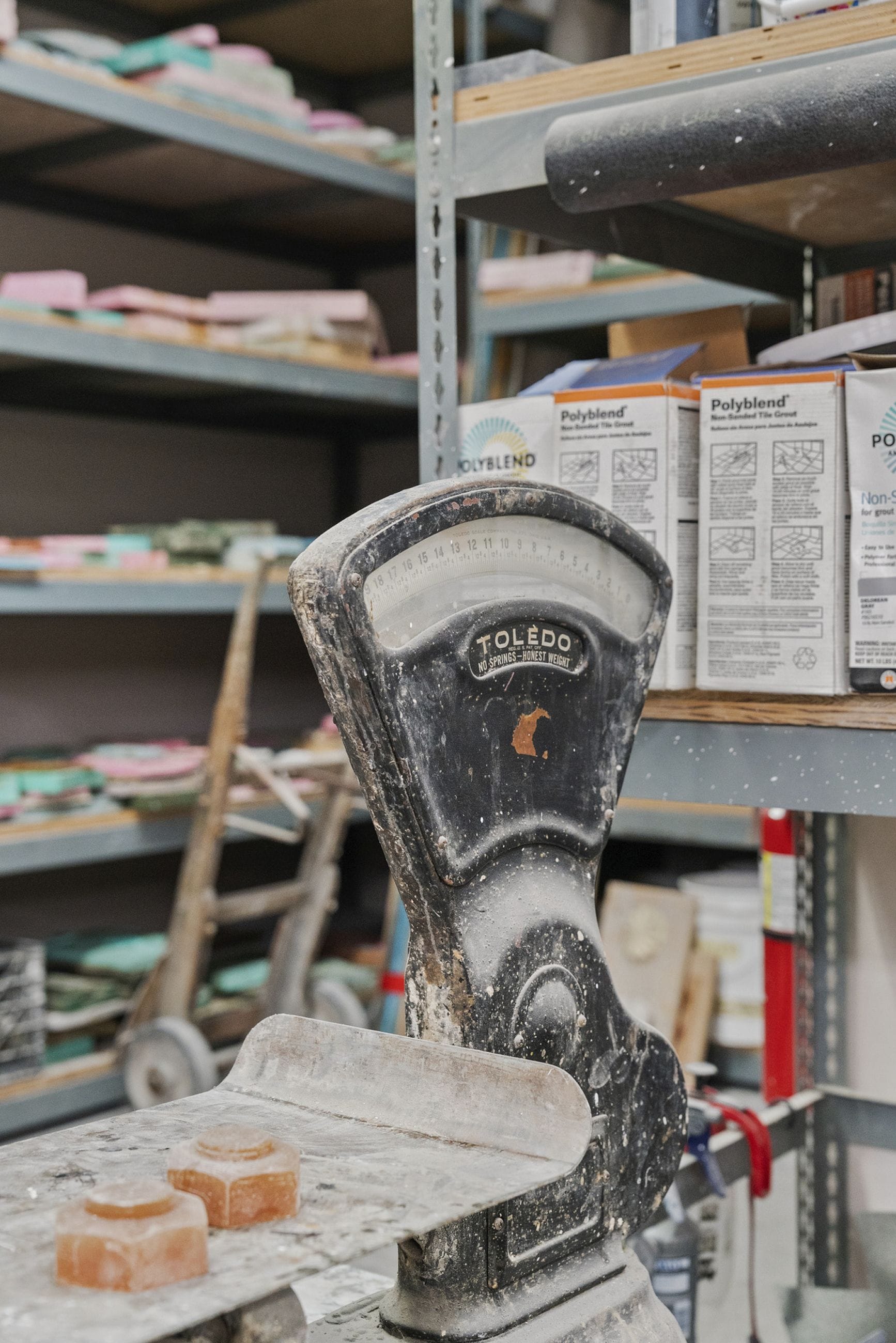 A dusty, black vintage Toledo scale with two orange hexagonal weights on its pan, in a workshop setting with shelves of supplies.