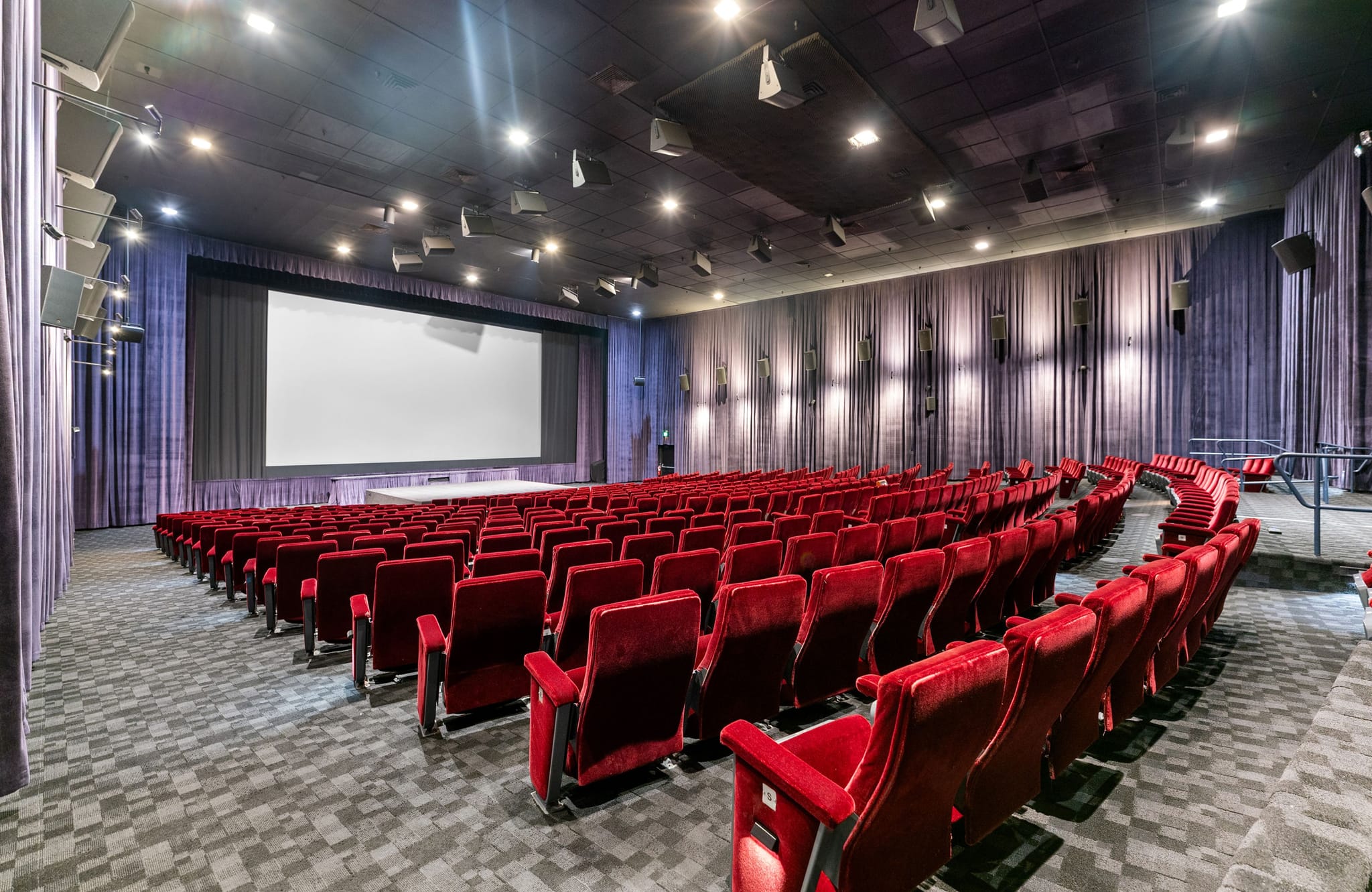An empty cinema hall with rows of red velvet seats facing a large white screen.