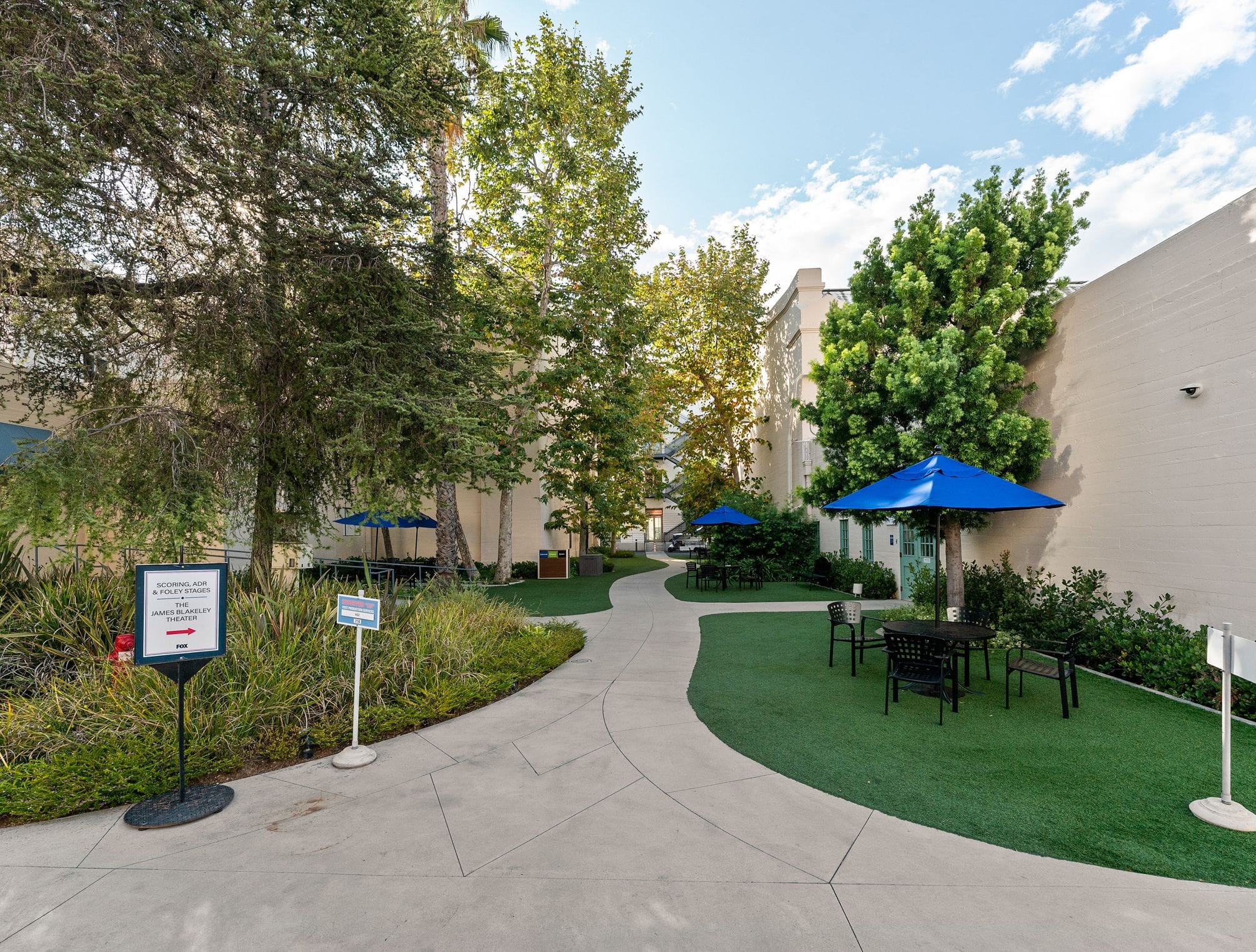 Outdoor patio with a winding concrete path, artificial grass, trees, and seating under blue umbrellas.