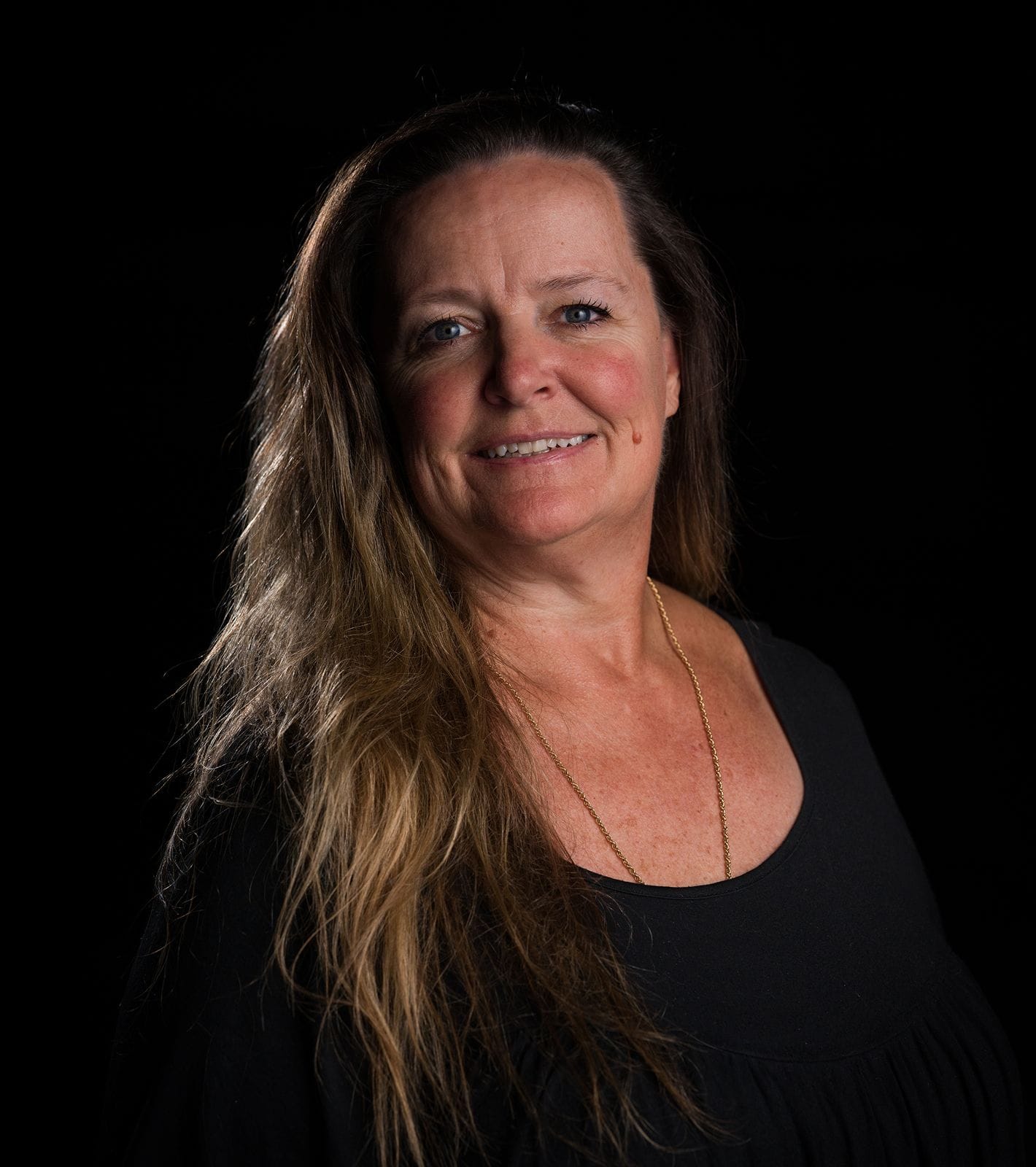 A smiling woman with long brown hair, wearing a black top and gold necklace, against a black background.