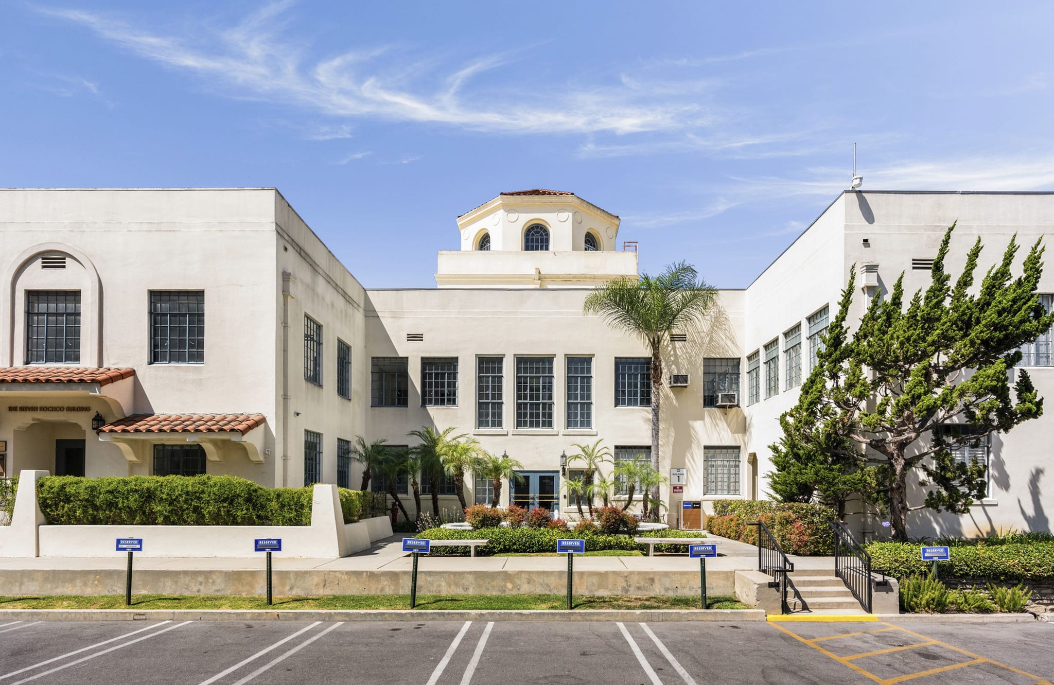 Exterior view of a cream-colored building with a central courtyard garden, topped by a domed cupola, and a parking lot in the foreground.