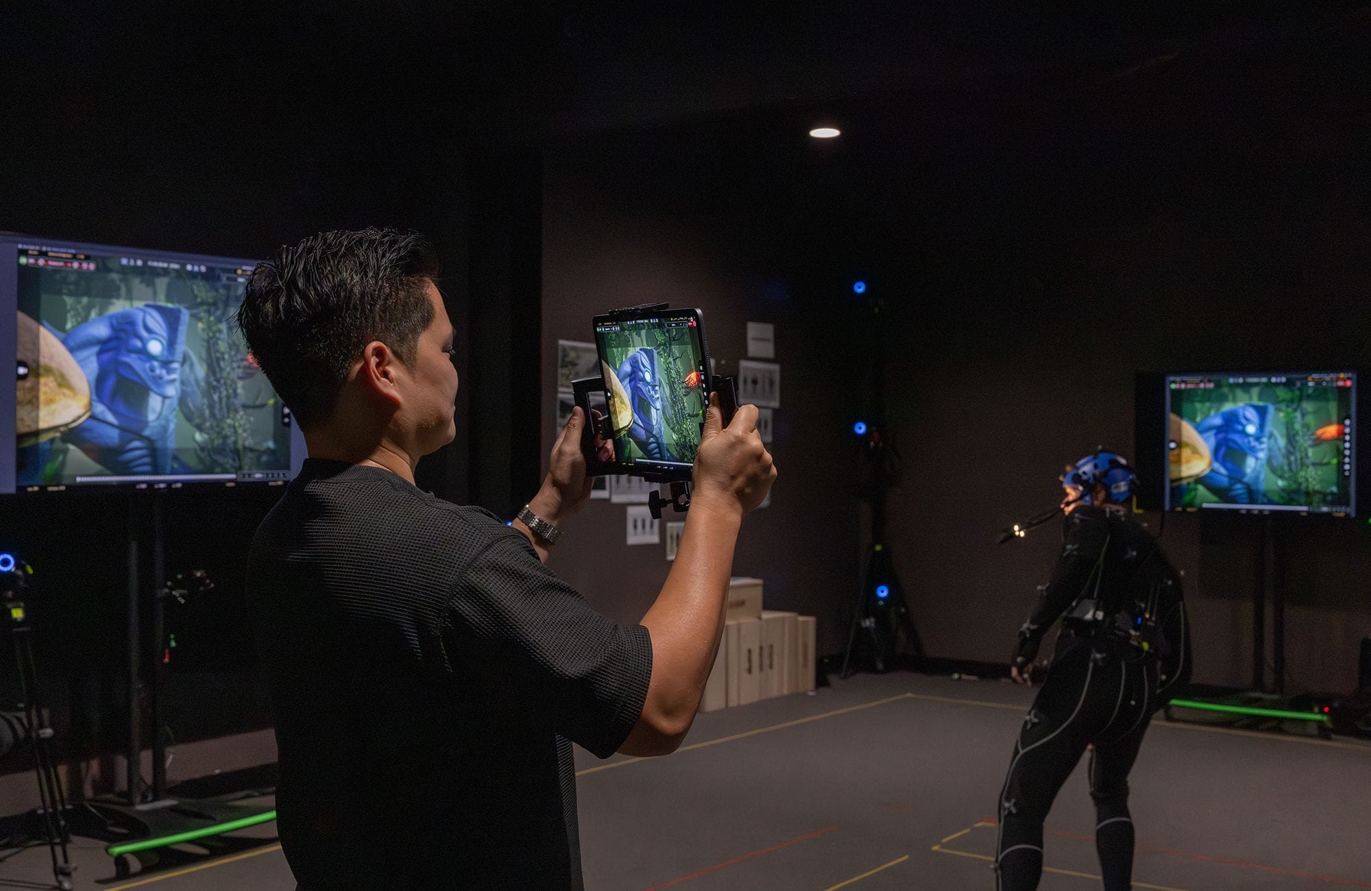 A man monitors a real-time virtual character on a tablet in a motion capture studio, with a performer in a suit.