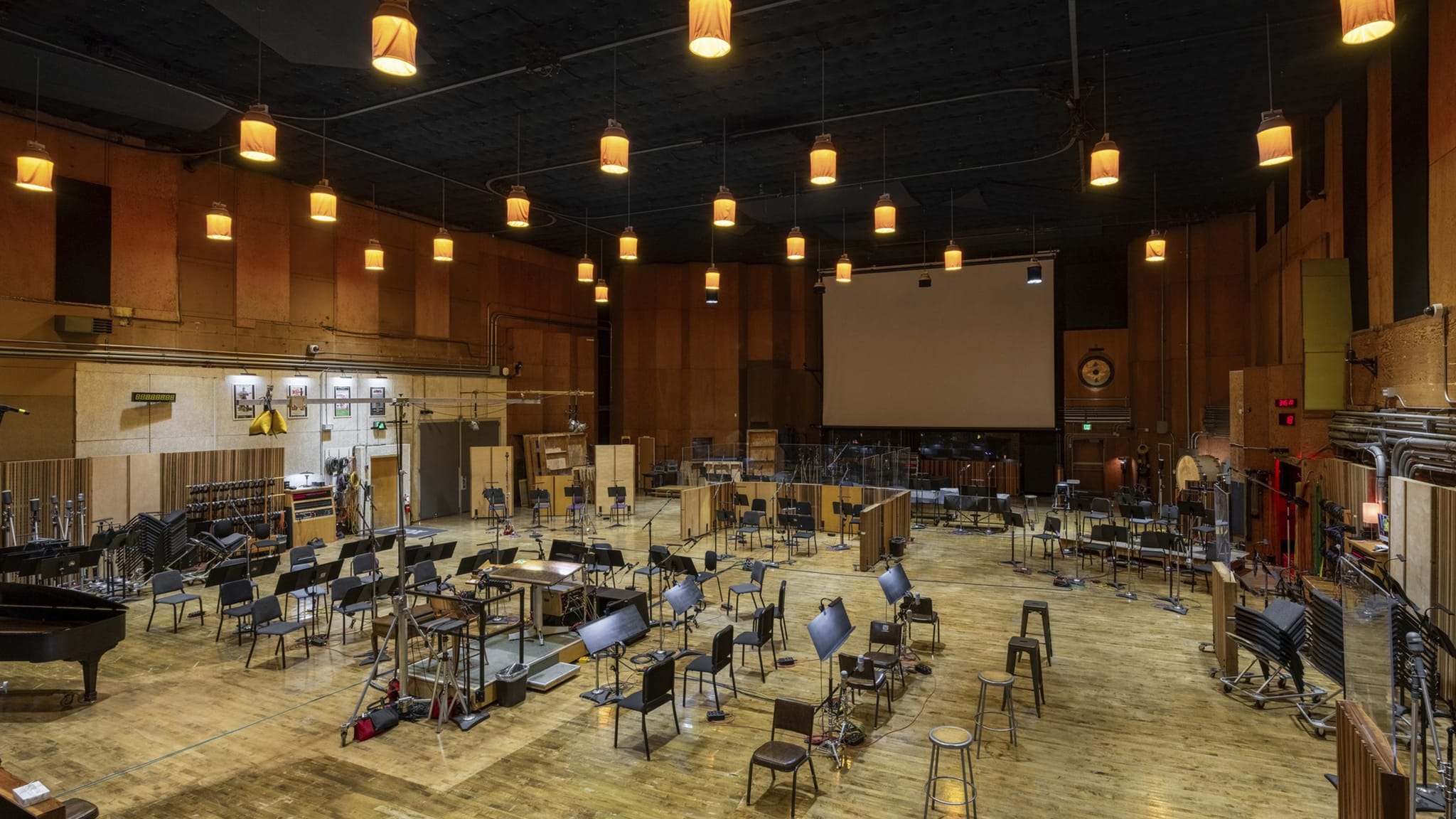 A large professional recording studio set up for an orchestra, with numerous chairs, music stands, a grand piano, and a large projection screen.
