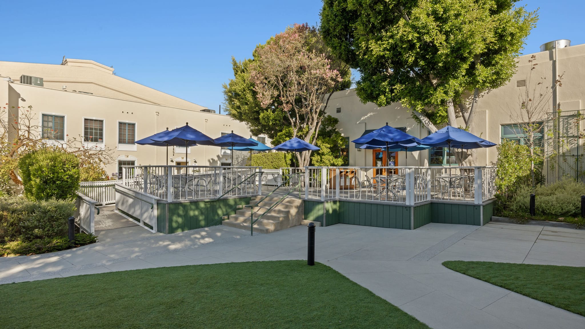 An outdoor patio with blue umbrellas, tables, and chairs, accessible by ramp and stairs, surrounded by buildings and trees.