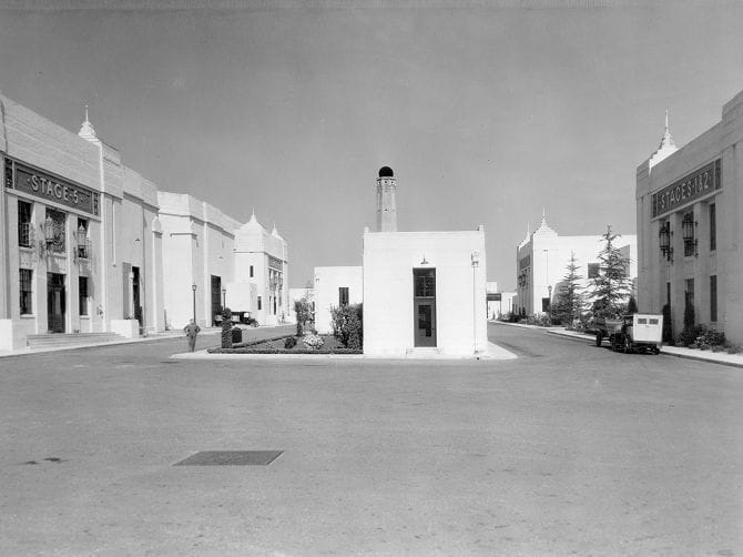 Vintage black and white photo of a studio lot street, showing large buildings "STAGE 5" and "STAGE 12", a central building with a chimney, and a classic car.