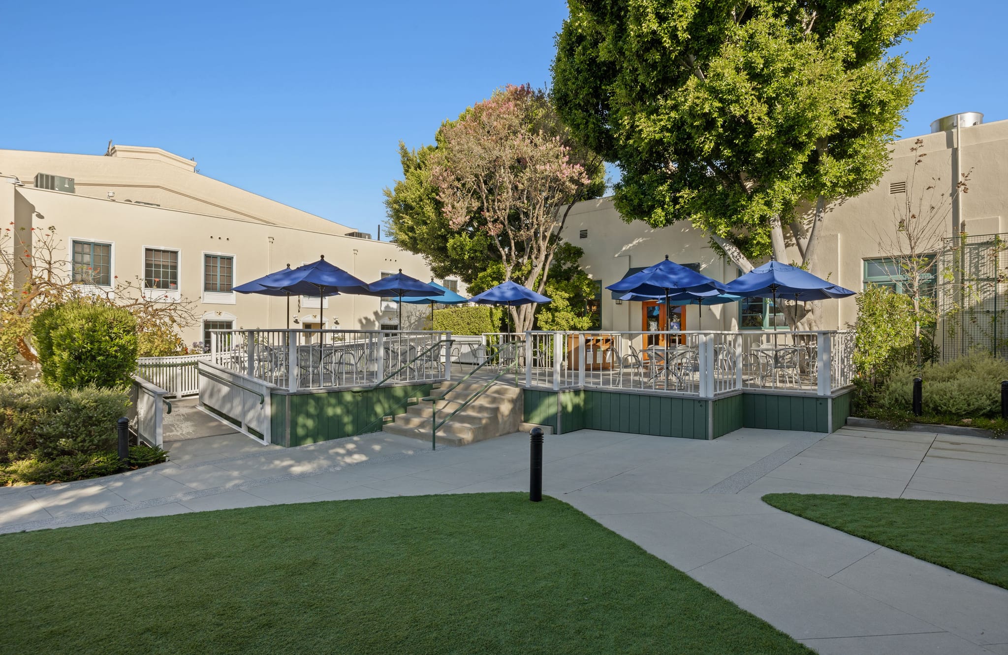 An outdoor patio with blue umbrellas, tables, and chairs, accessed by a ramp and stairs from a grassy area, with a building in the background.