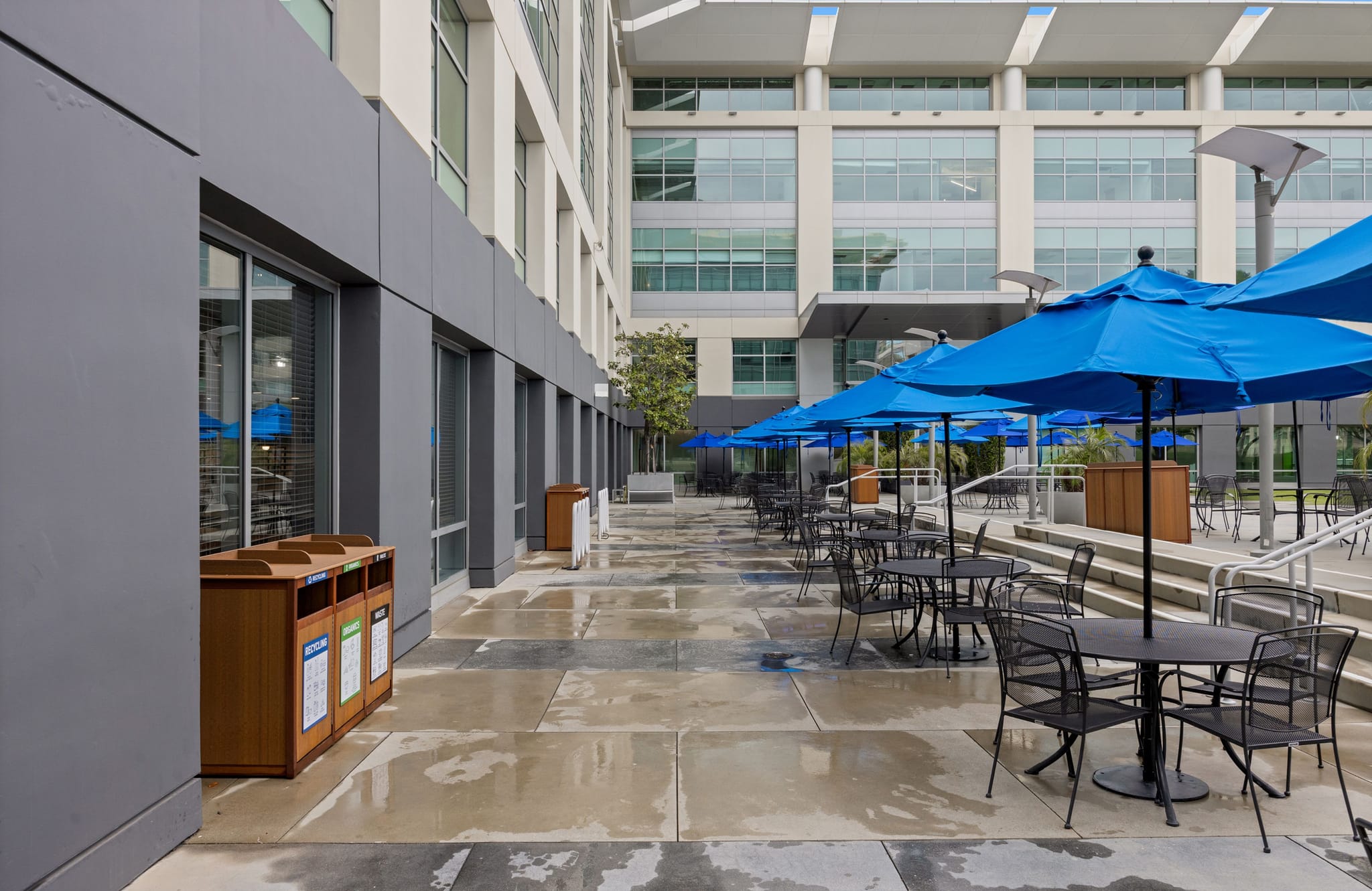 A long outdoor patio with black tables, chairs, and bright blue umbrellas in front of a modern office building.