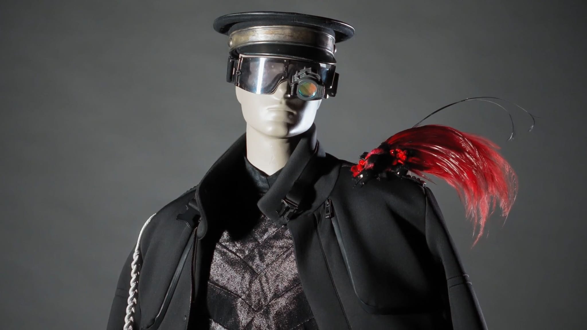 Mannequin wearing a black military-style uniform with futuristic silver visor glasses, a red feathered epaulet, and a grey textured shirt.