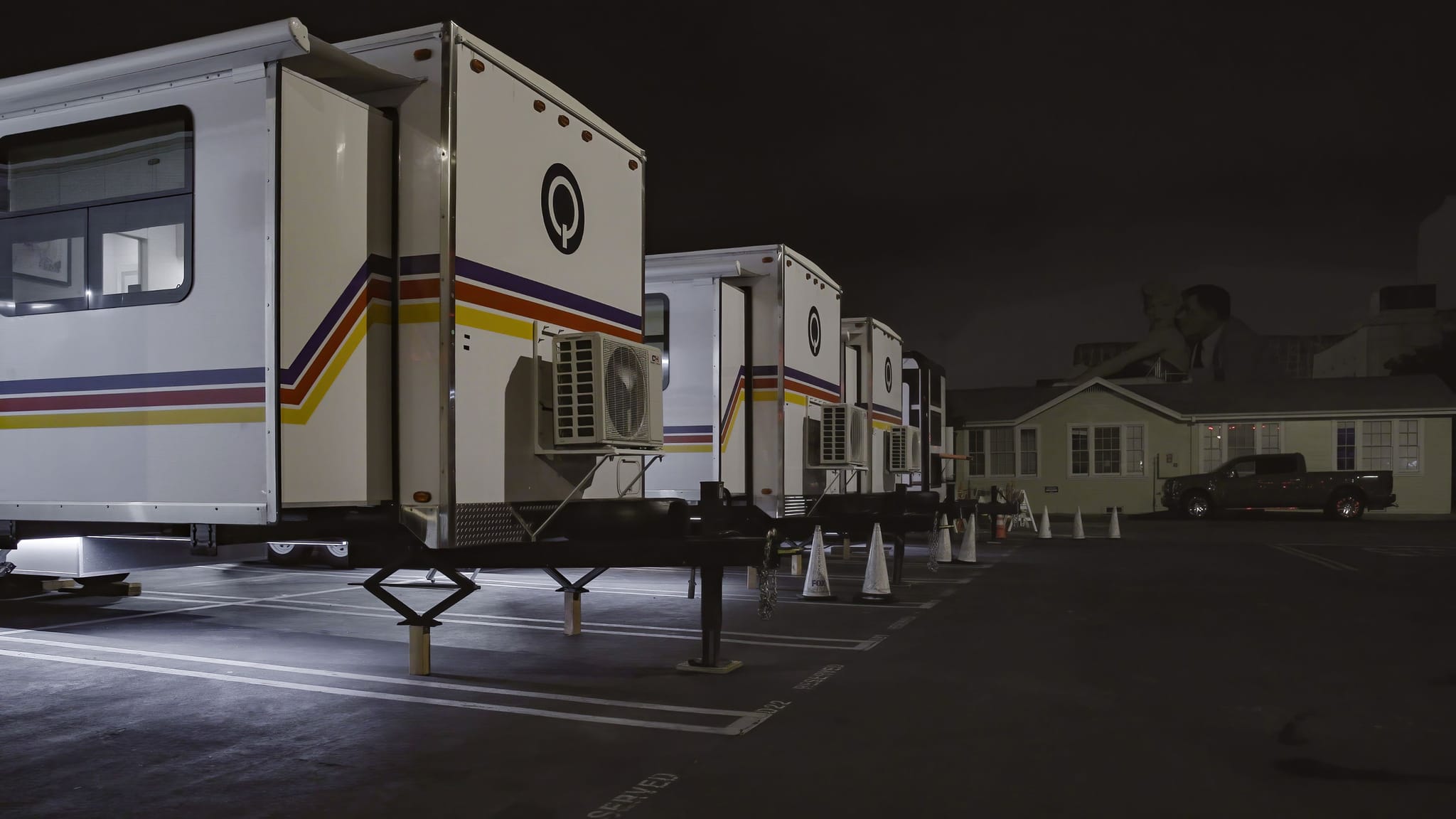 A line of white production trailers with colorful stripes parked at night, with a faint, giant human silhouette visible above a building in the background.