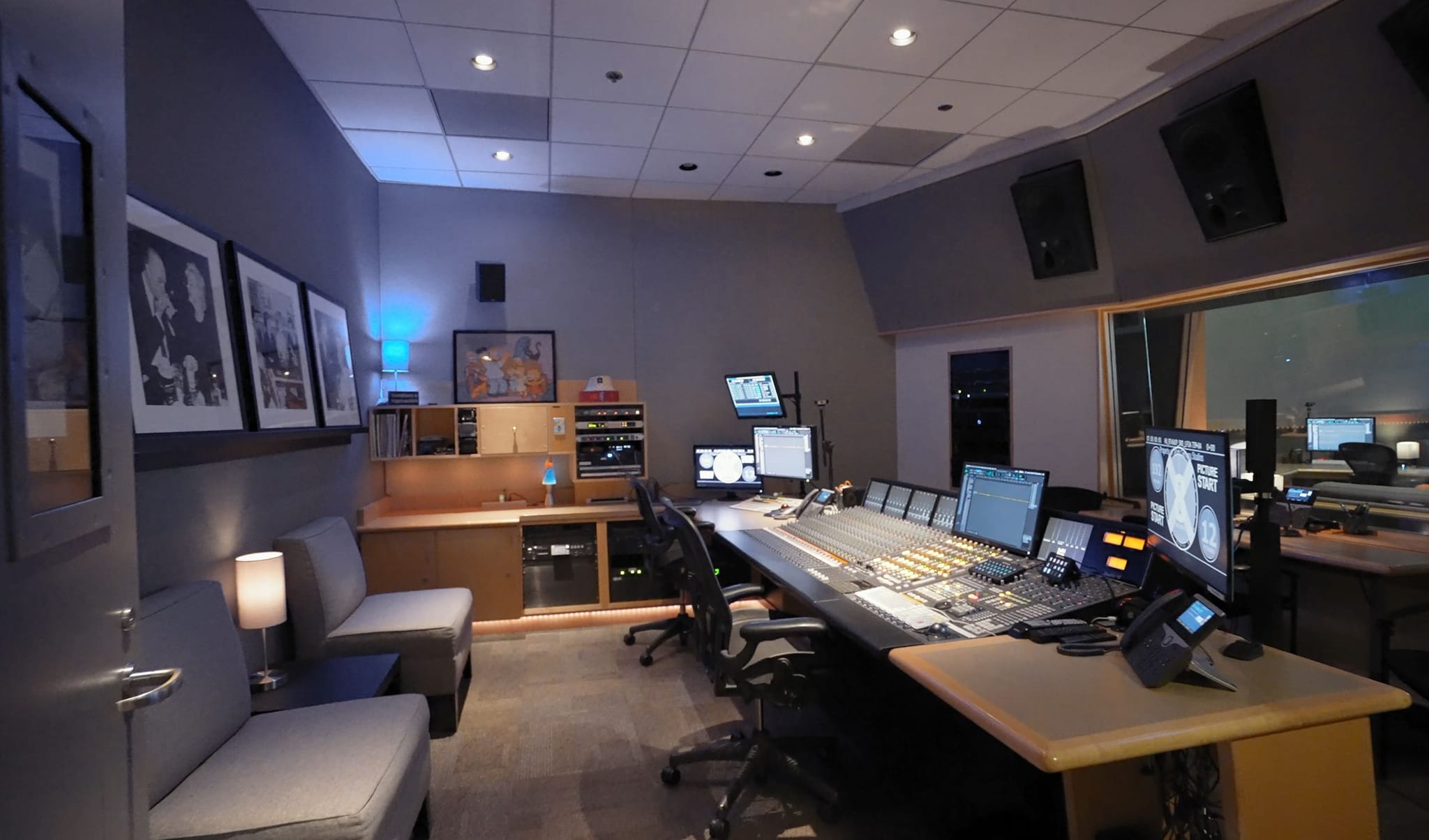 A recording studio control room with a large mixing console, multiple monitors, and a seating area.