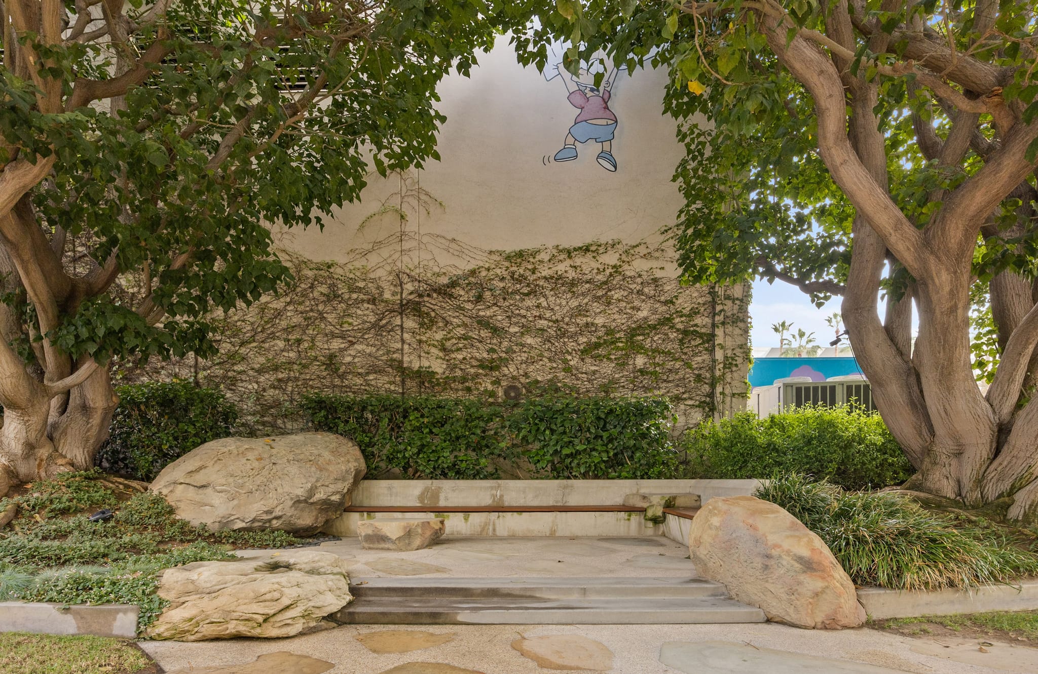 A landscaped area with large trees and boulders flanking a path, leading to a vine-covered wall with a mural of a swinging child.
