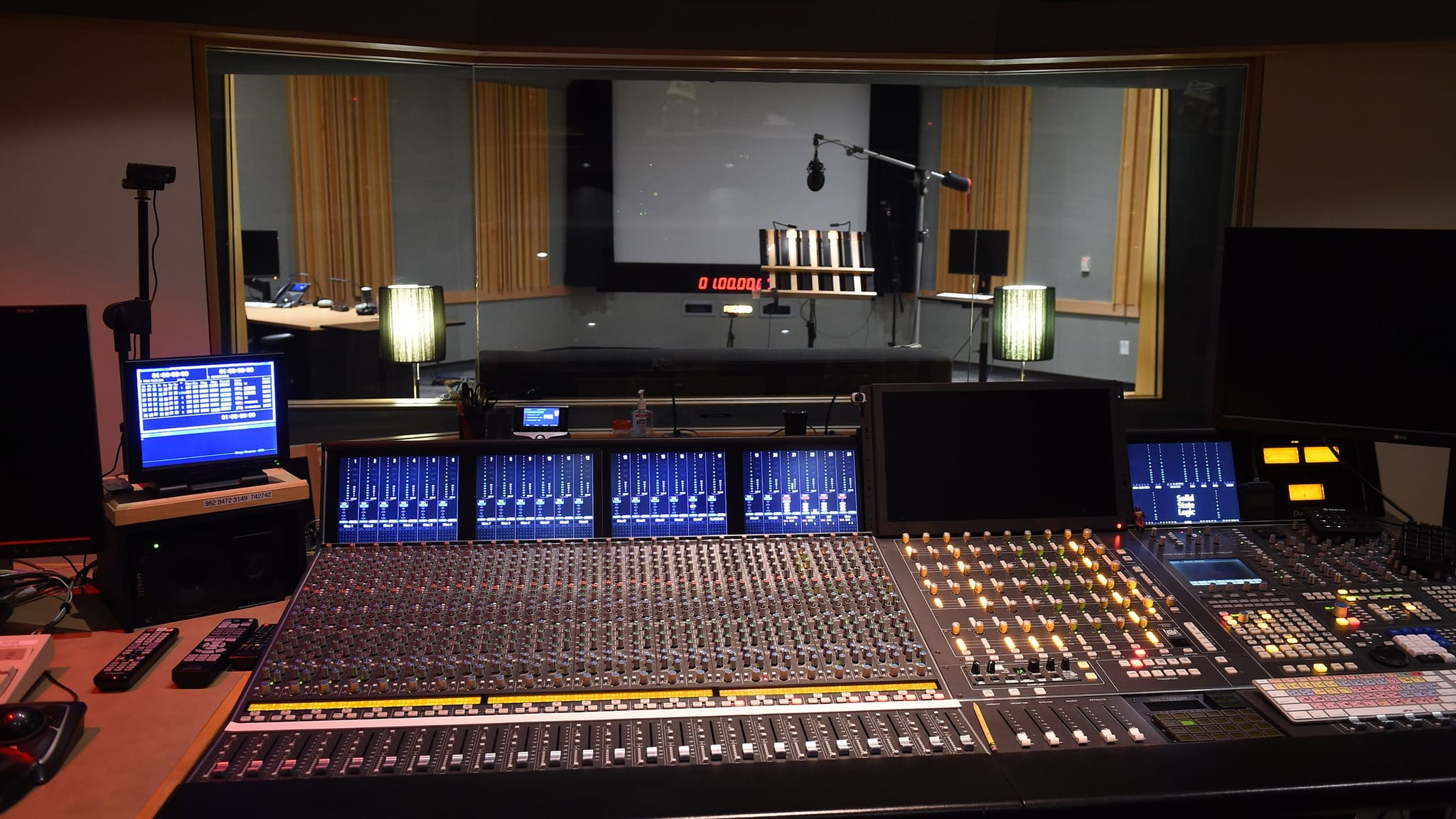 A recording studio control room with a large mixing console in the foreground, looking into a recording booth with a microphone.