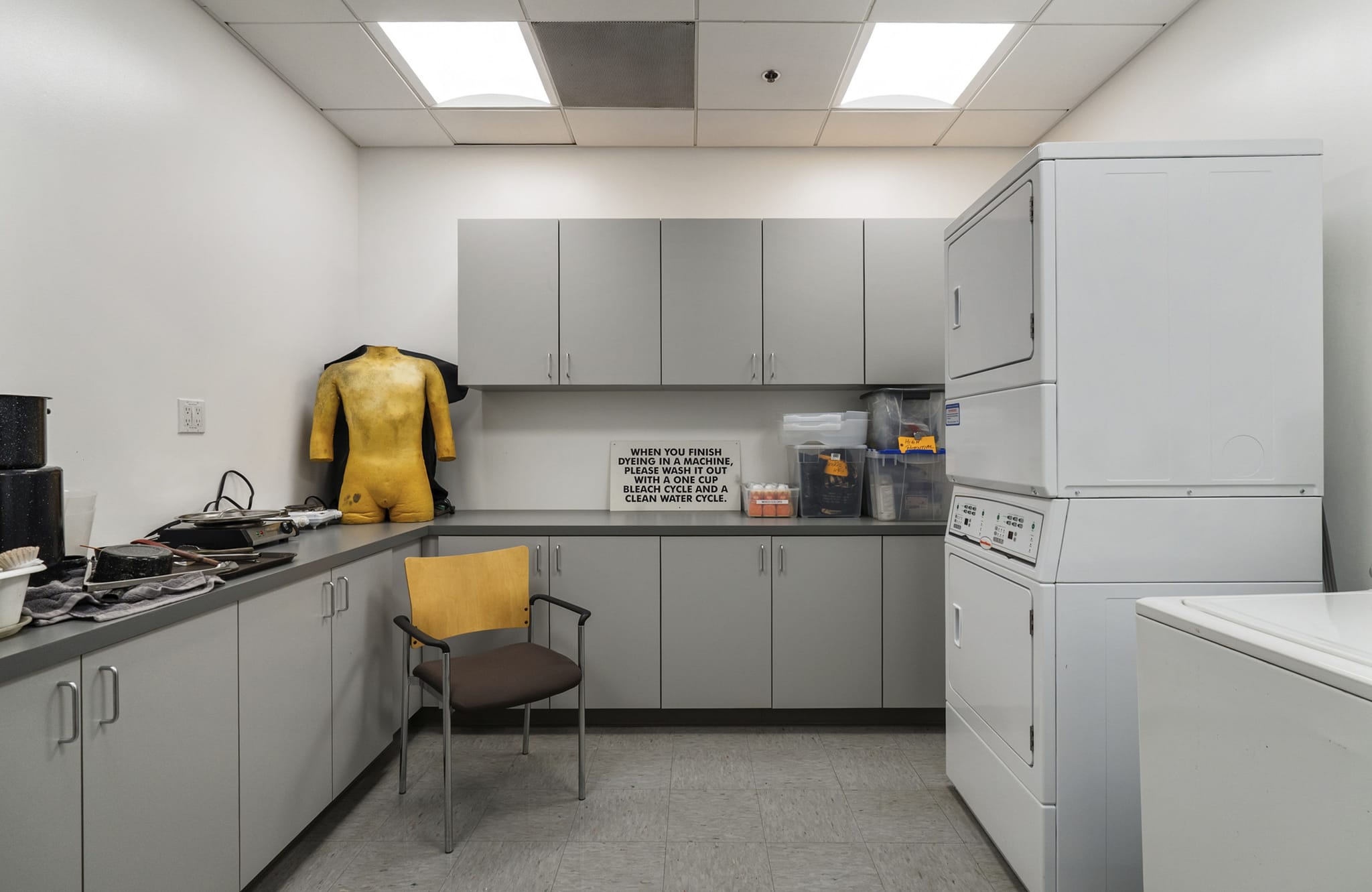 A functional room with grey cabinets, a headless yellow mannequin, and stacked white laundry machines. A sign on the back wall gives dyeing instructions.