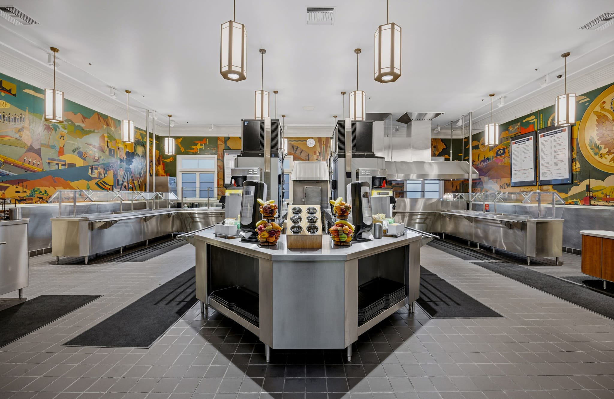 An empty cafeteria with colorful murals on the walls, stainless steel buffet lines, and a central island counter.