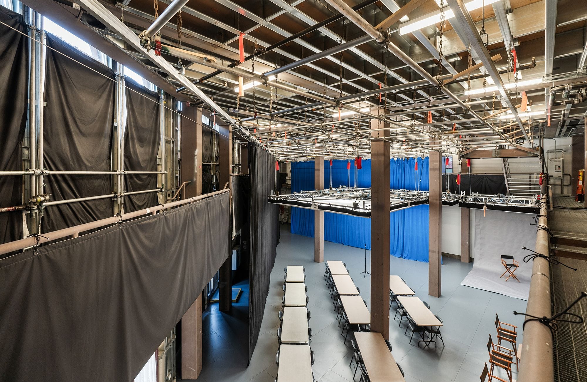 A large, versatile studio space with high ceilings, a balcony, rows of tables, blue and white backdrops, and a director's chair.