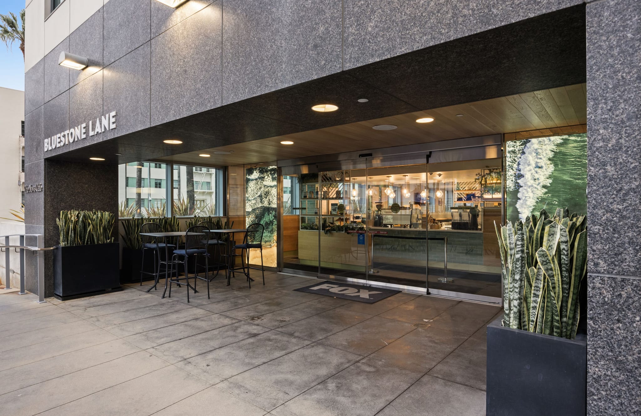 Exterior of Bluestone Lane cafe in a modern dark stone building, with outdoor seating and potted plants.