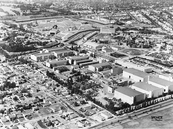 Vintage black and white aerial view of a large film studio lot amidst a city and residential neighborhoods.