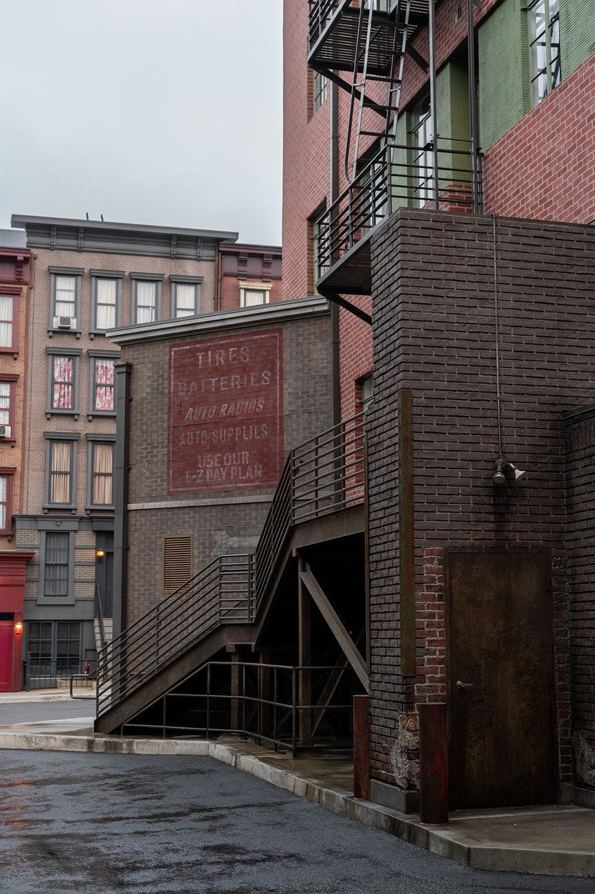 Urban alleyway with brick buildings, a metal staircase, and an old painted auto supplies ad on a wall.
