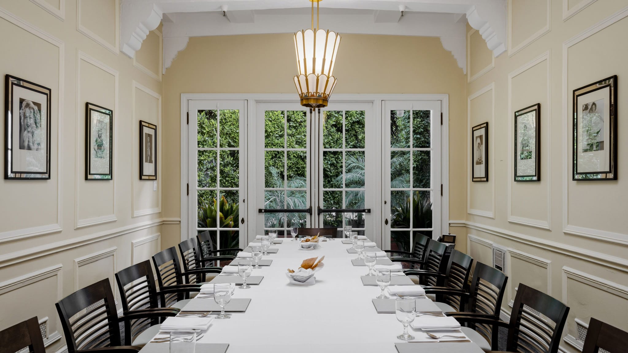 A long dining table set for many in a cream-walled room with framed art, a chandelier, and French doors to a garden.