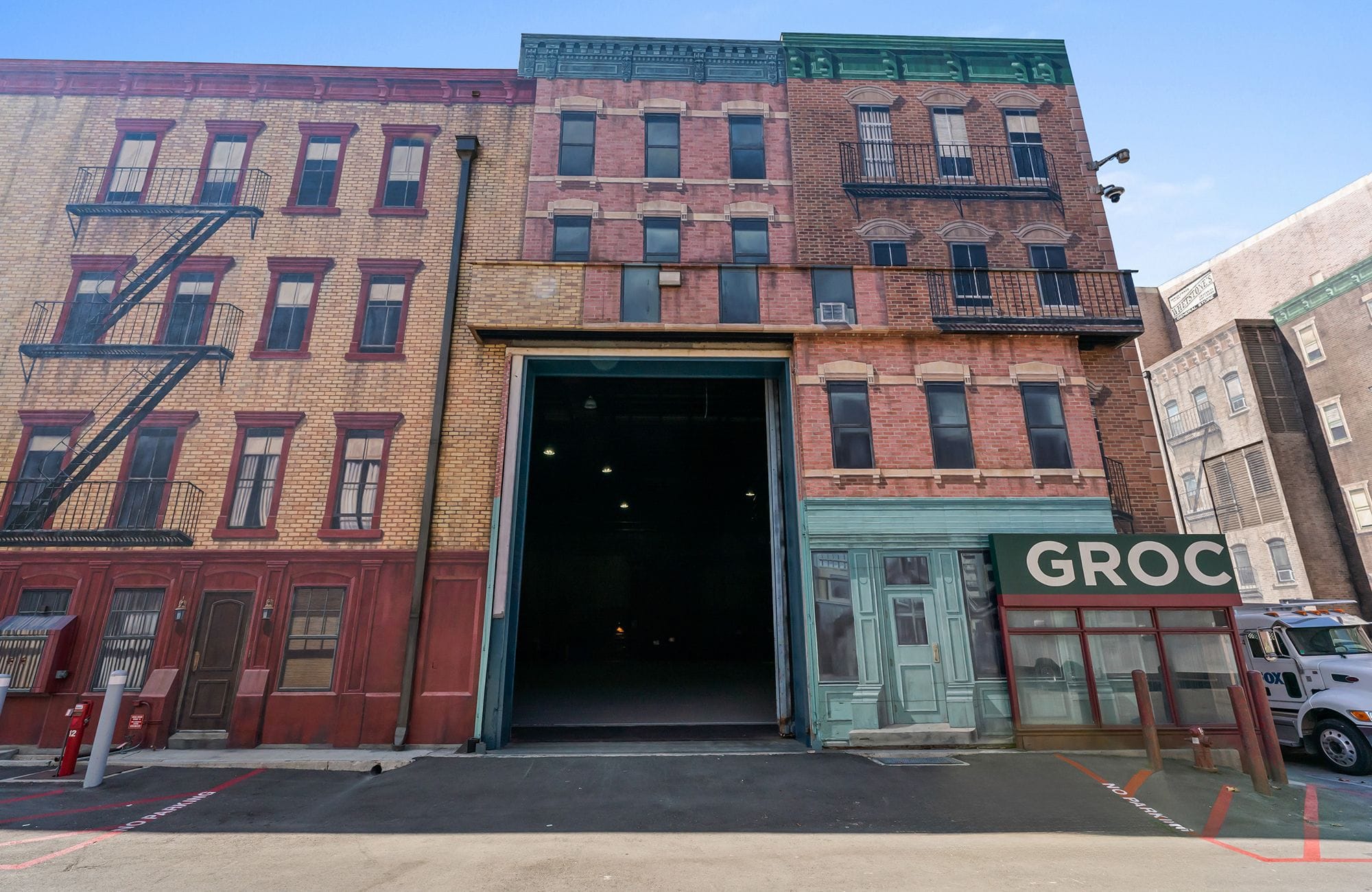 Urban facade featuring brick buildings with fire escapes and a 'GROC' sign, with a large open bay revealing a dark interior.