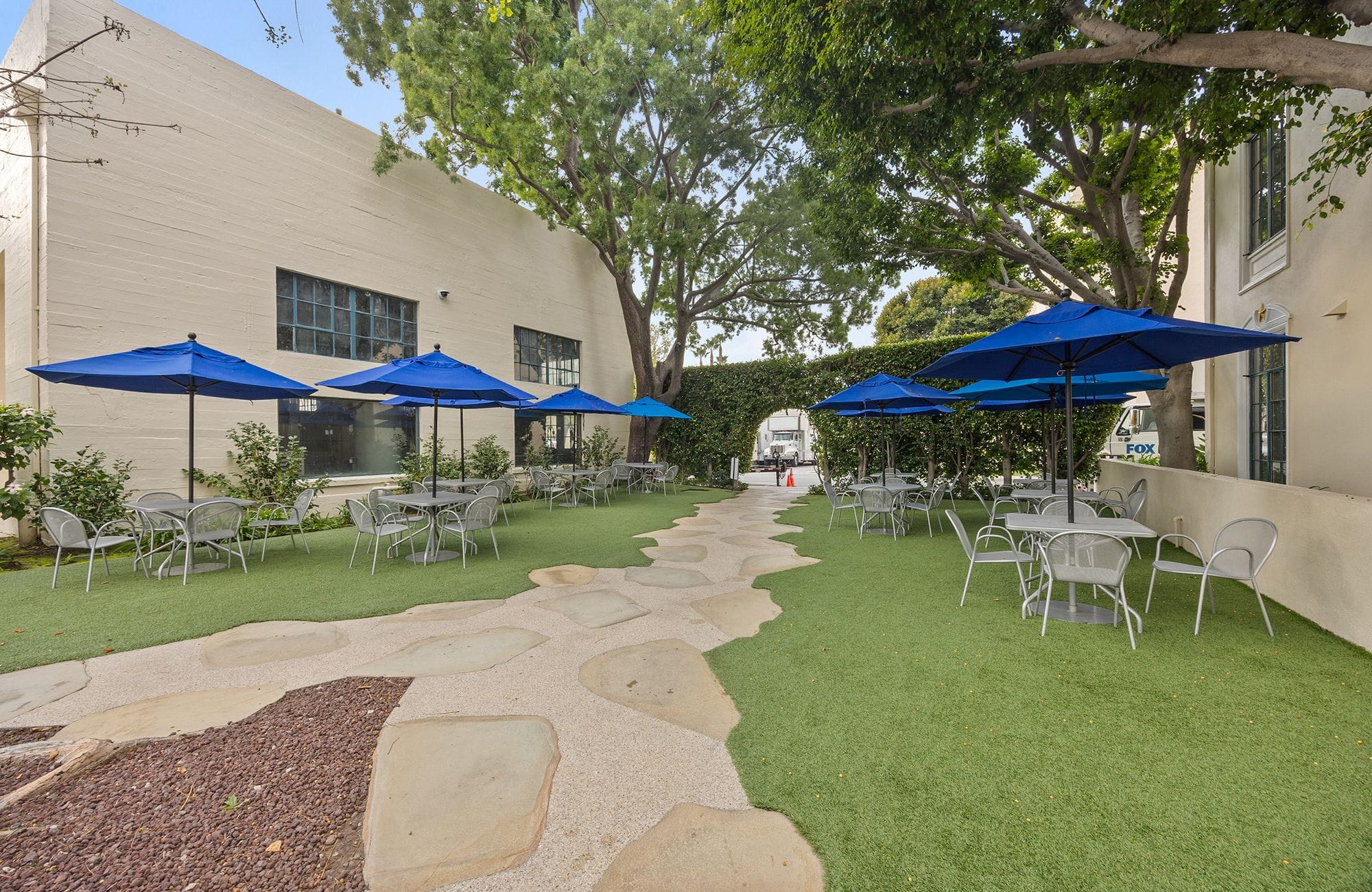 Outdoor patio with tables, chairs, blue umbrellas, and a stone pathway on artificial grass.