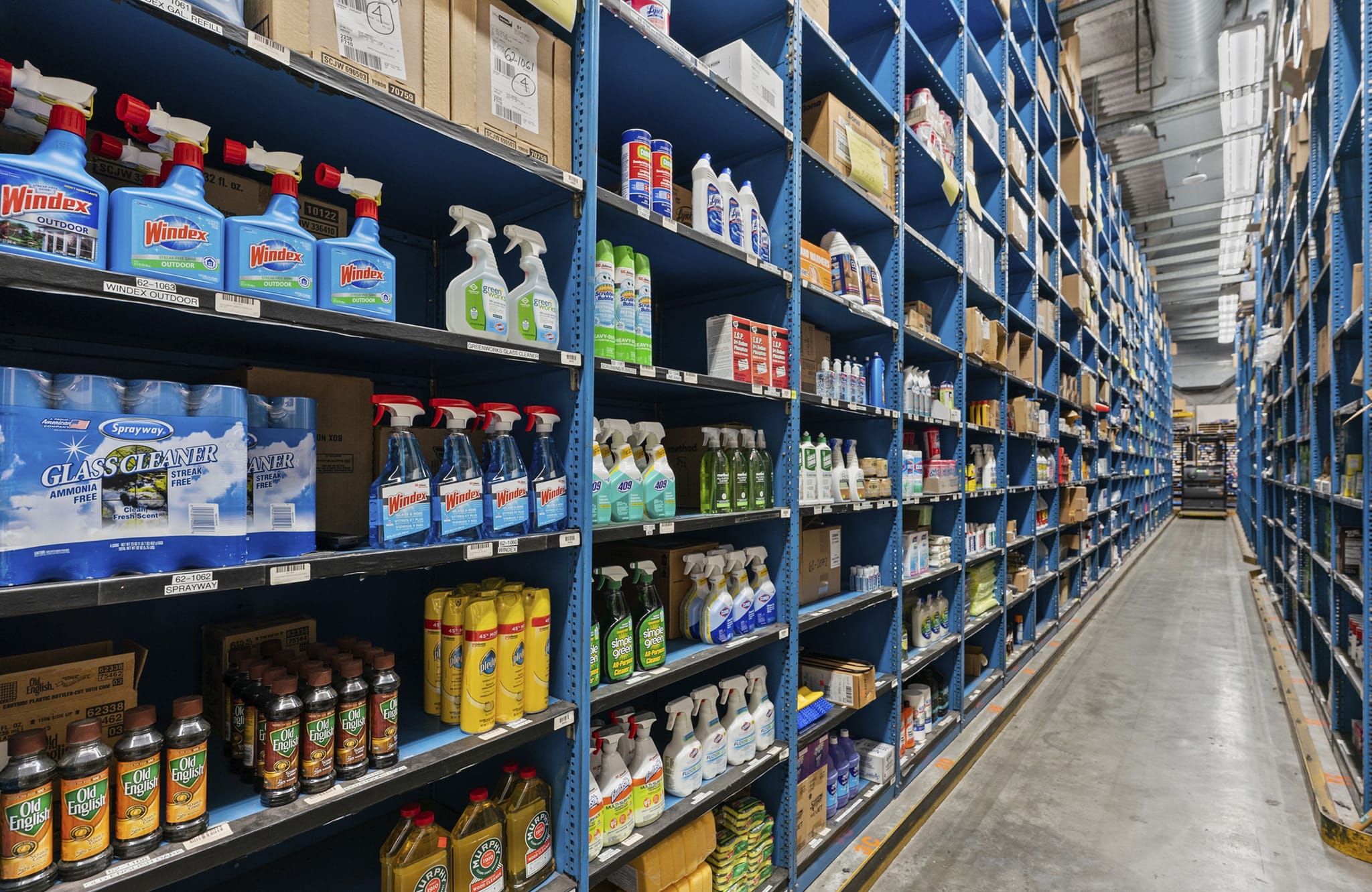 A long warehouse aisle with tall blue shelves filled with various products, predominantly cleaning supplies.