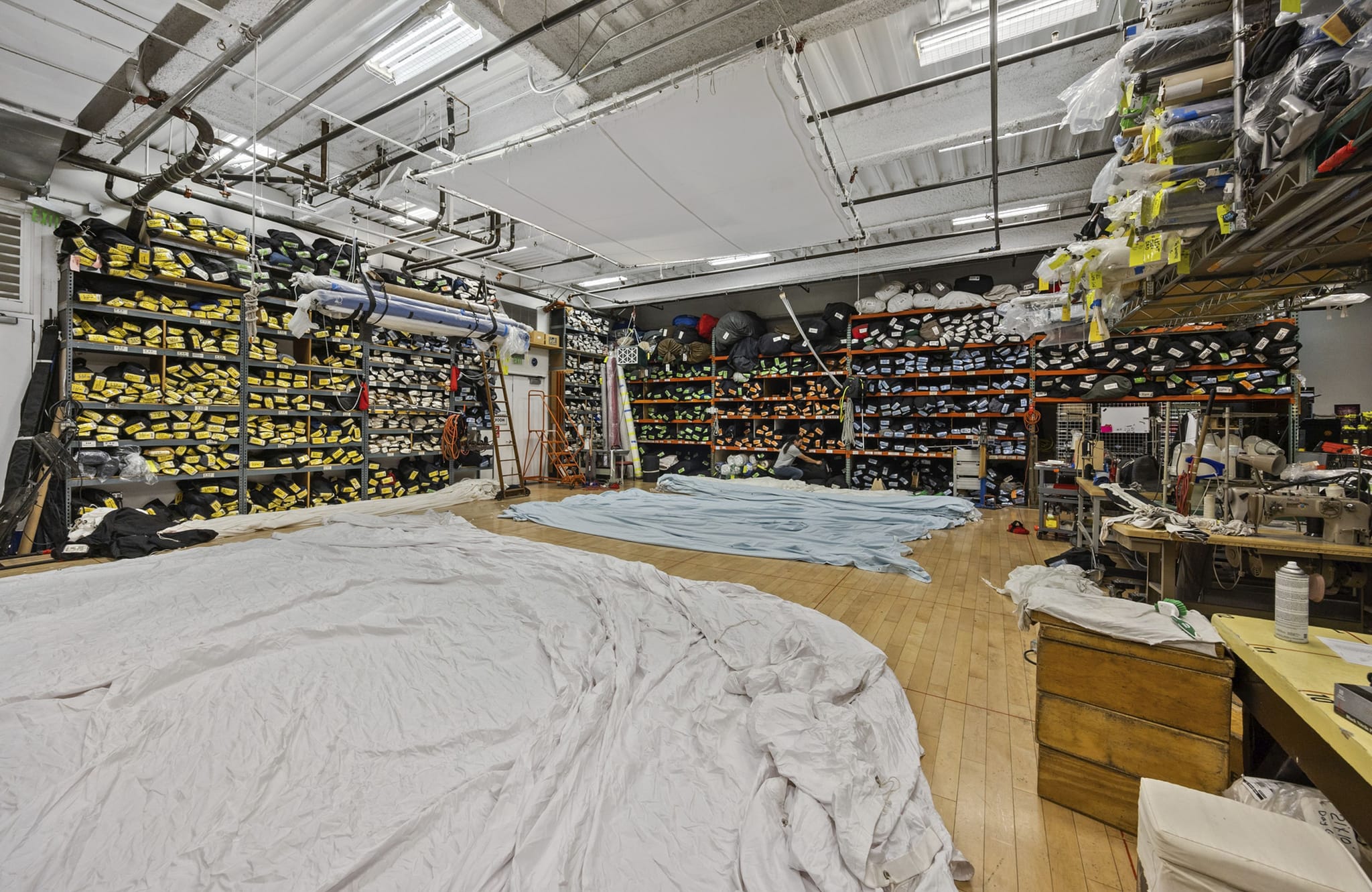 A large workshop with white fabric spread on a wooden floor, surrounded by shelves of fabric rolls and sewing machines.