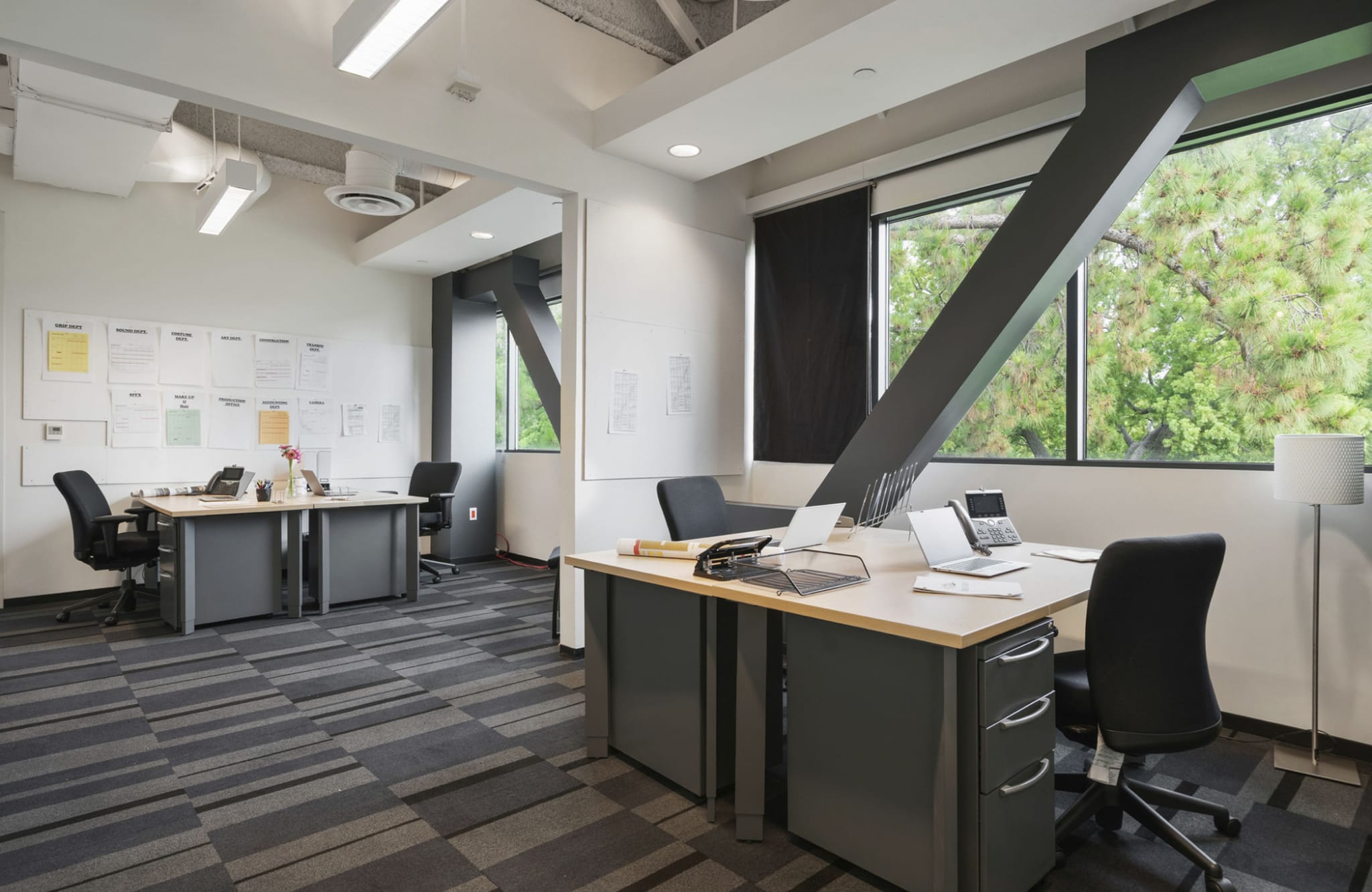A modern open-plan office with multiple workstations, a striped carpet, and large windows revealing green trees and a prominent diagonal structural beam.