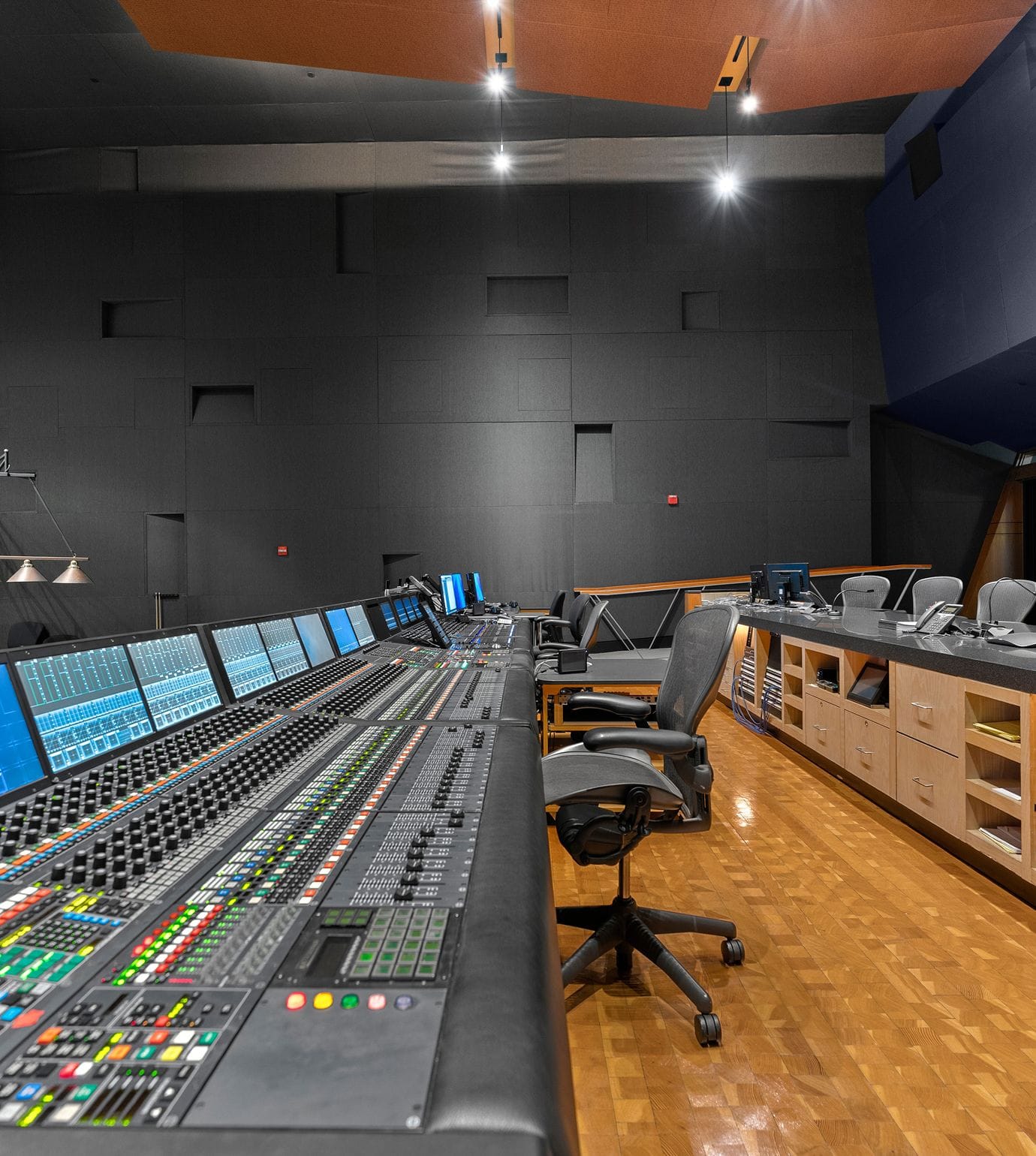 A dark, modern media studio with a large projection screen, extensive multi-monitor control desk, and a long conference table.