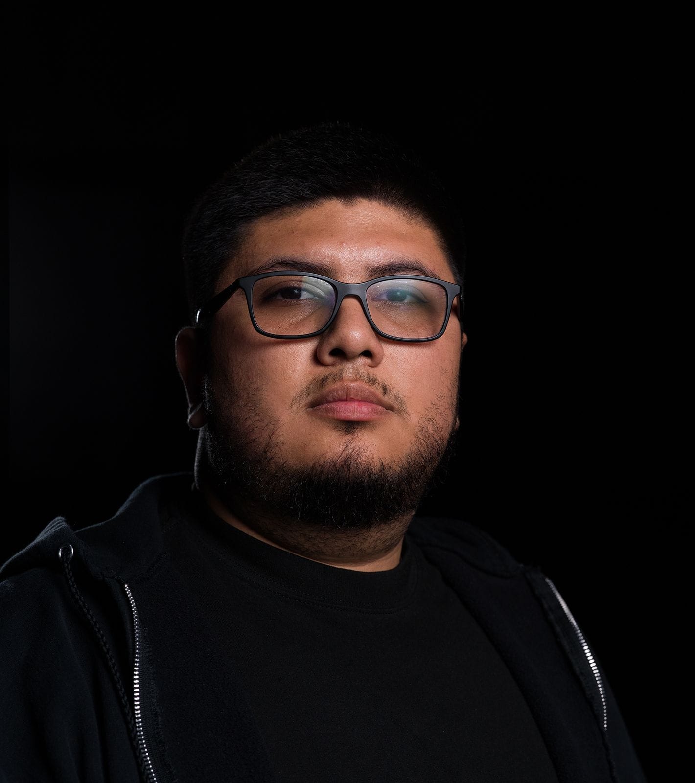 A man with dark hair, a beard, and glasses looks directly at the camera, wearing a black hoodie against a dark background.