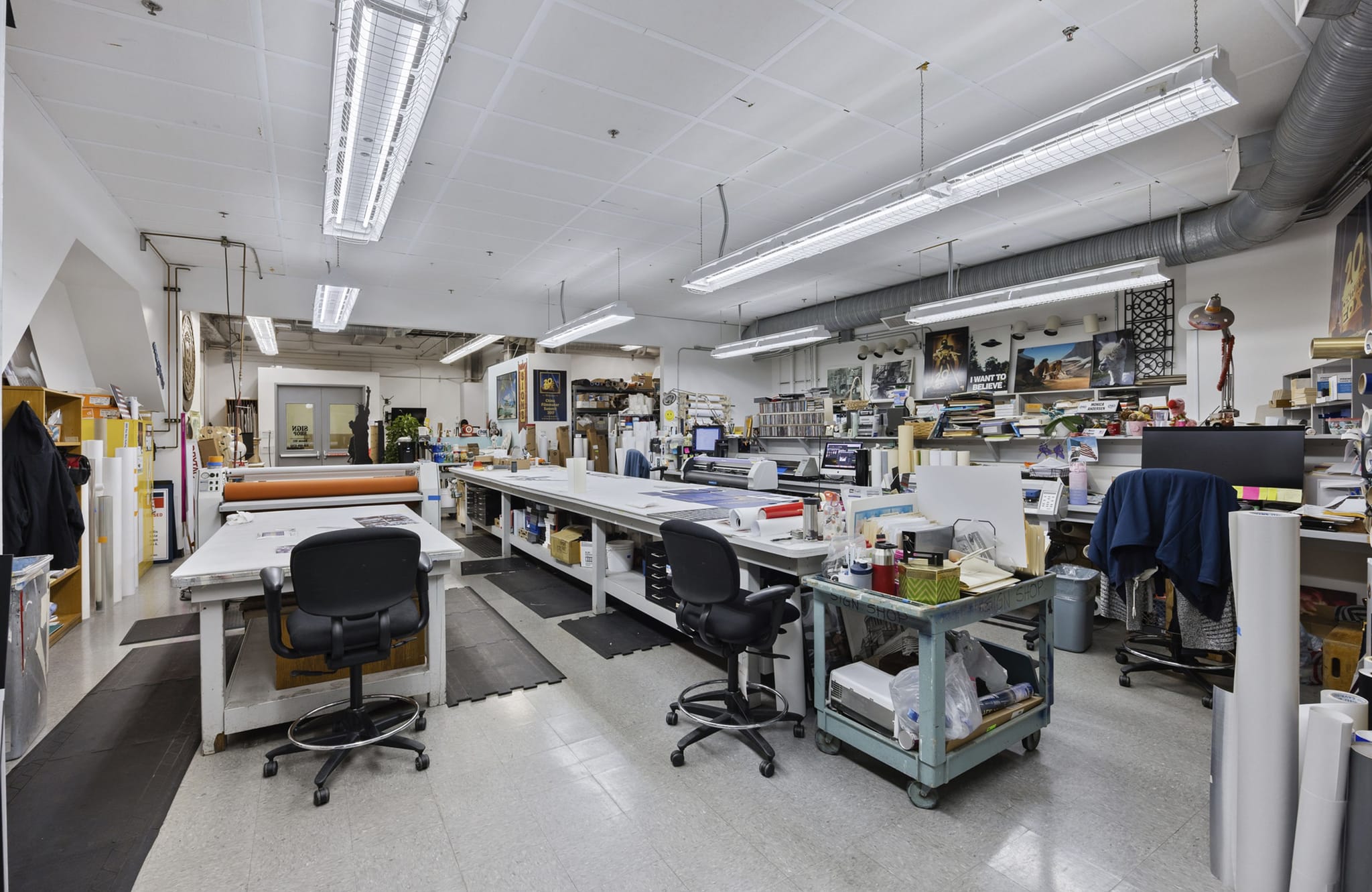 A brightly lit print production studio with long work tables, printing equipment, and various supplies.