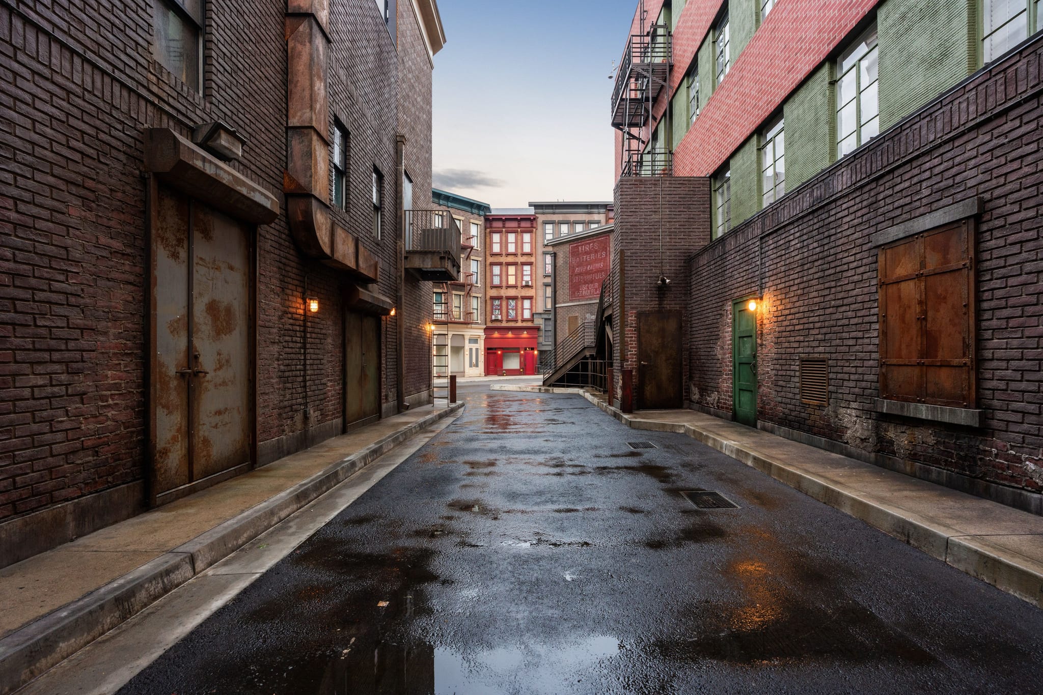 A wet alleyway between old brick buildings, with puddles on the pavement and more buildings visible in the distance.