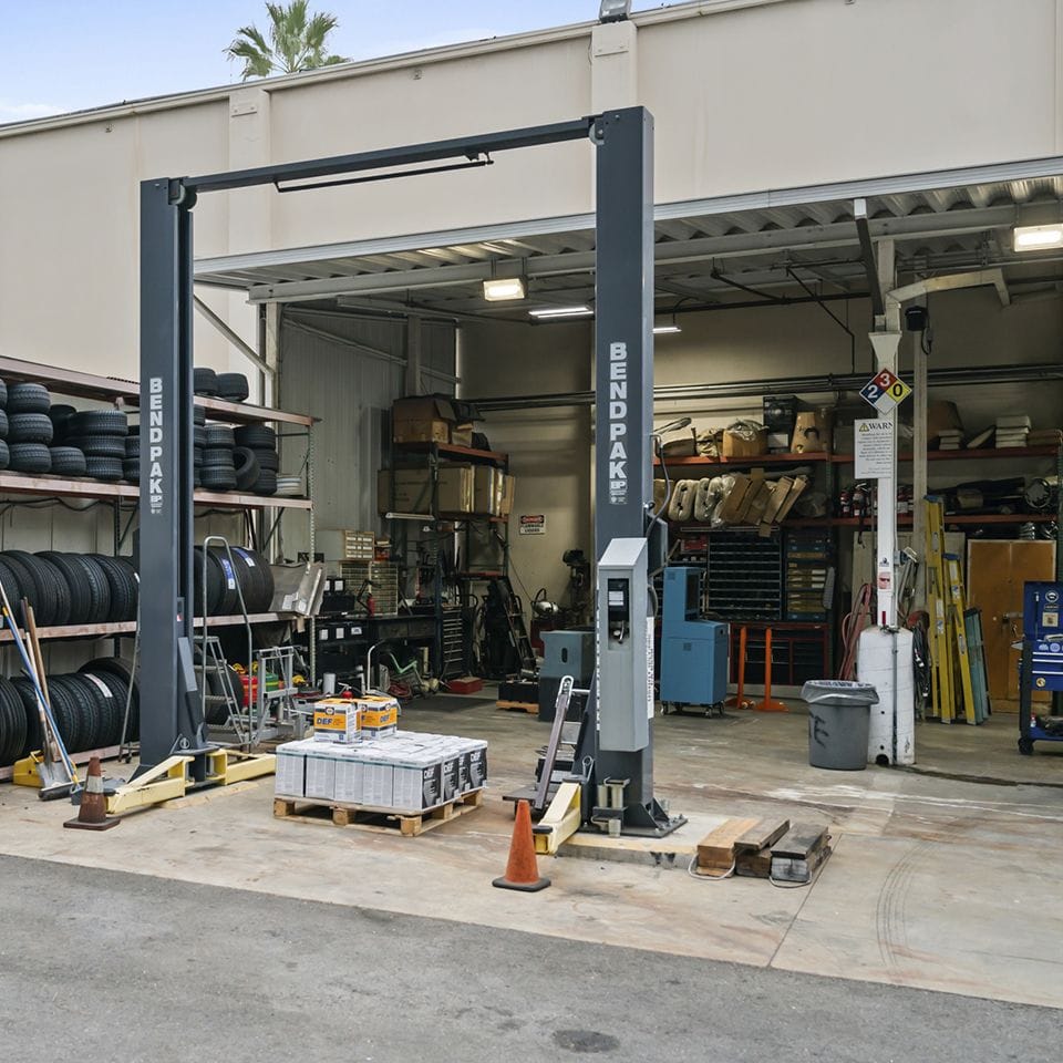 A two-post vehicle lift stands empty in a cluttered auto repair garage with shelves of tires and parts.