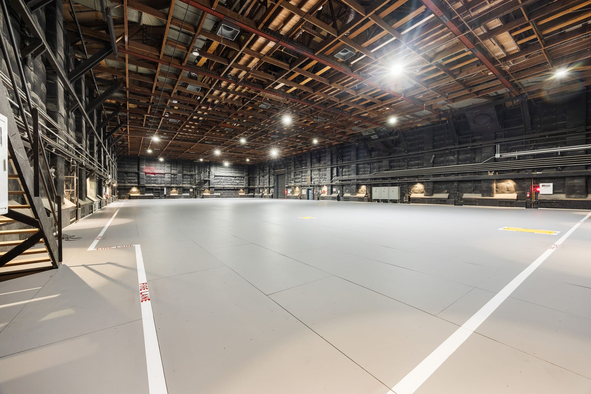 View of a large, vacant studio space featuring a high, exposed wood ceiling, dark walls, and a light grey floor with white lines.