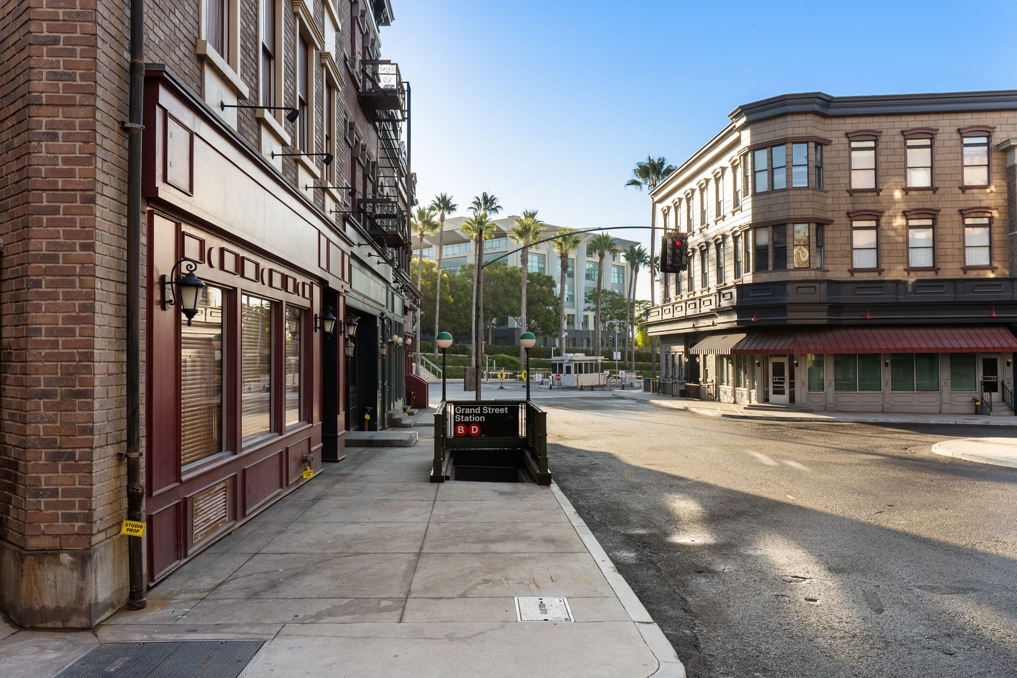 A quiet street corner with traditional brick buildings, a "Grand Street Station" subway entrance, and palm trees in the distance.