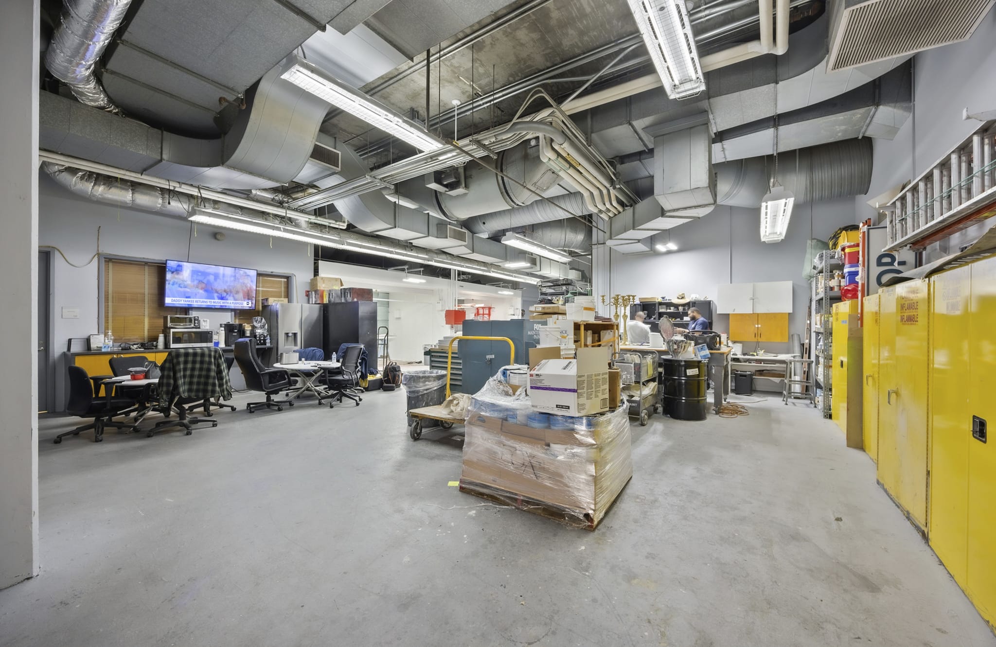 Large industrial space with exposed ceiling ducts, an office area, and yellow storage cabinets.