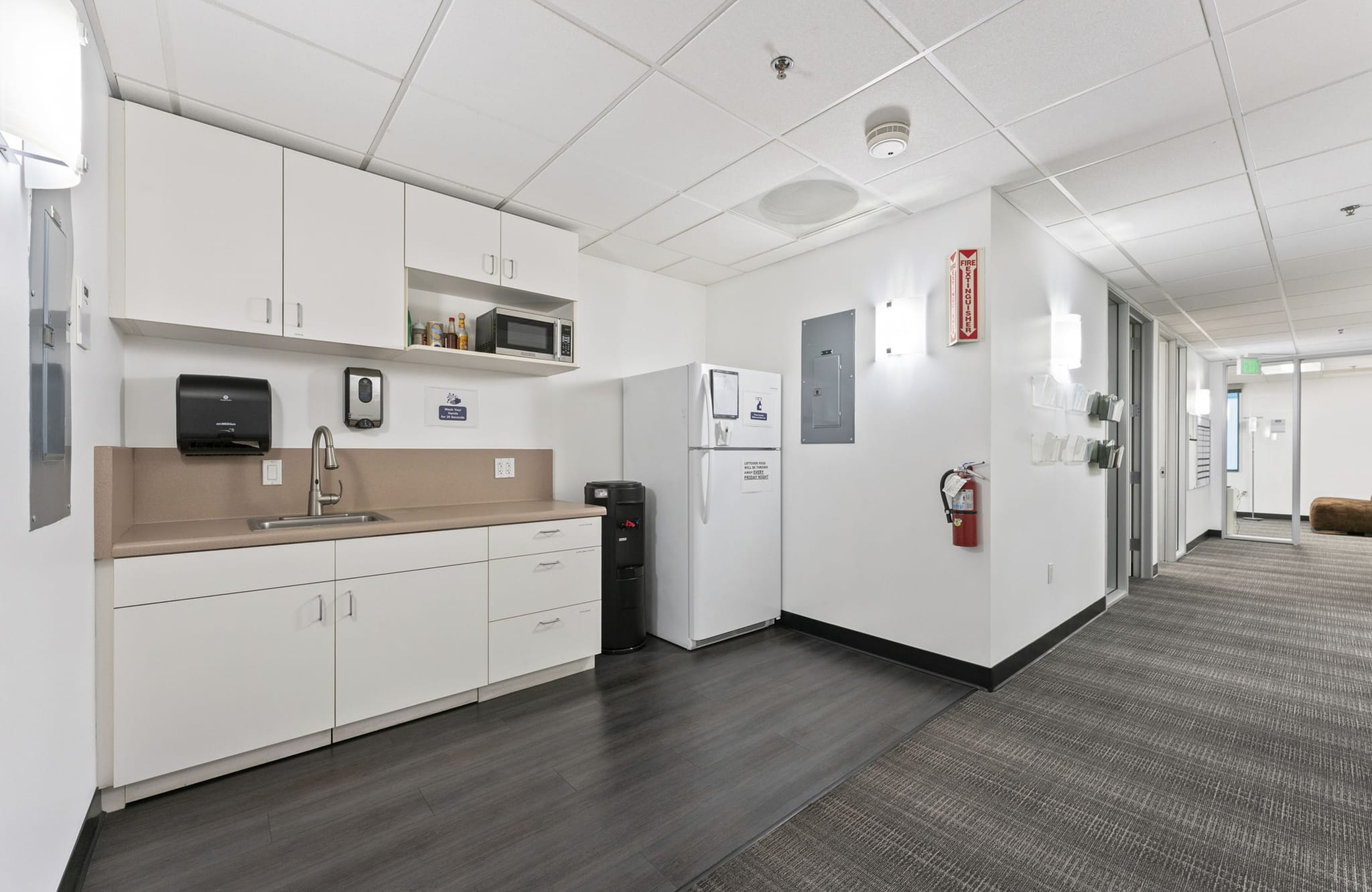 Office breakroom with white cabinets, sink, microwave, refrigerator, and water cooler, adjacent to a carpeted hallway.