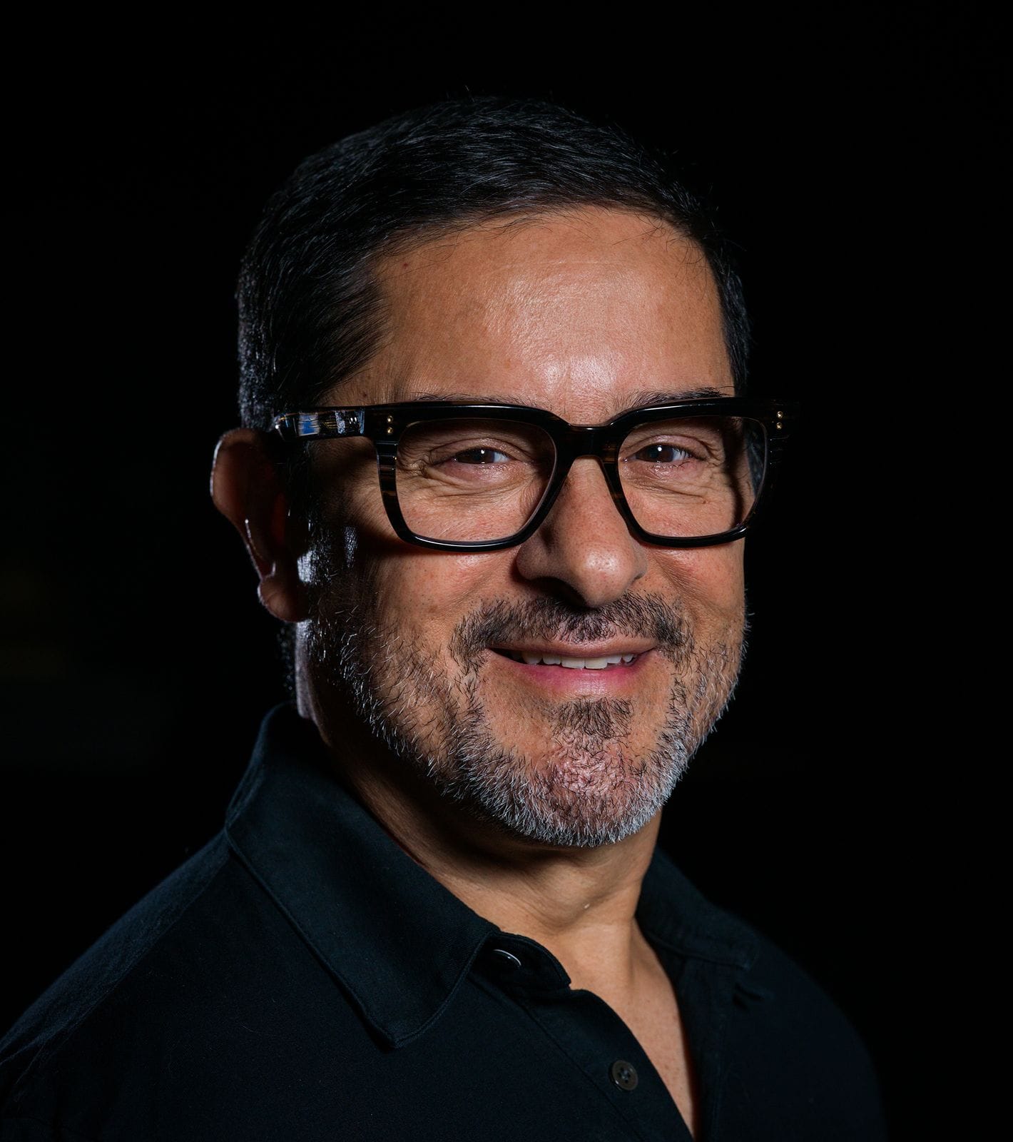 A smiling man with dark hair, a salt-and-pepper beard, and large black glasses against a dark background.