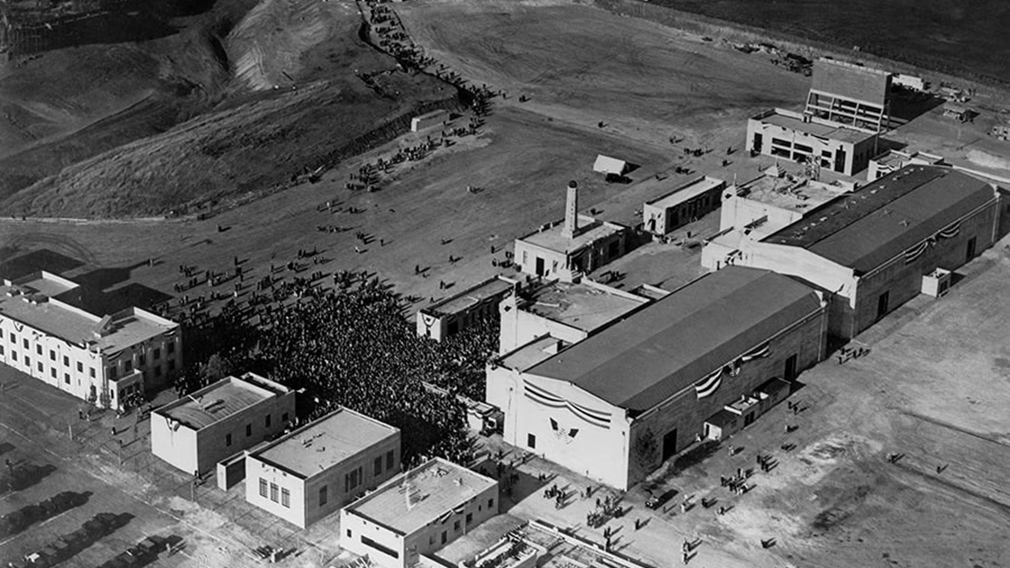 Aerial black and white photo of a film studio complex with a large crowd gathered outdoors, against a backdrop of hills.